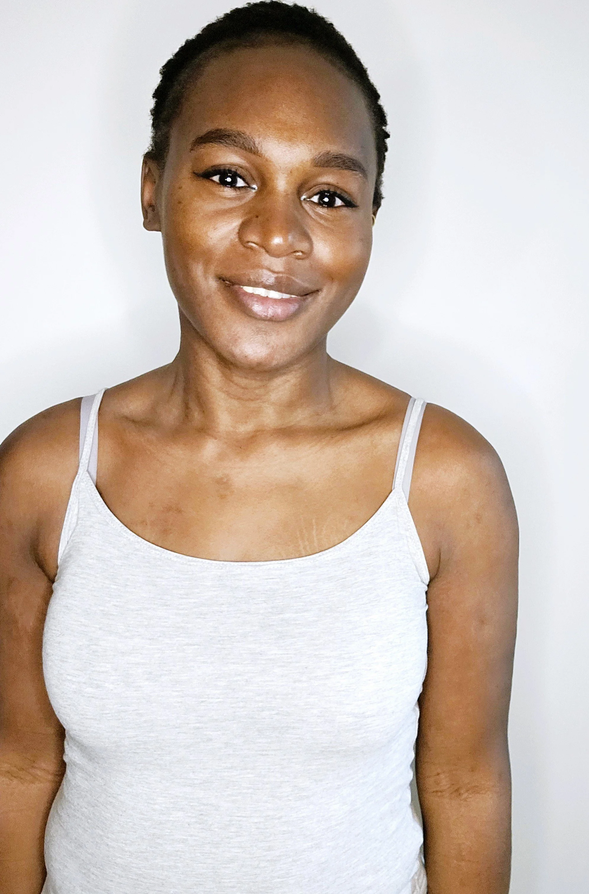 Portrait d'une femme souriante avec une peau brune et portant un débardeur gris clair, debout contre un fond blanc.