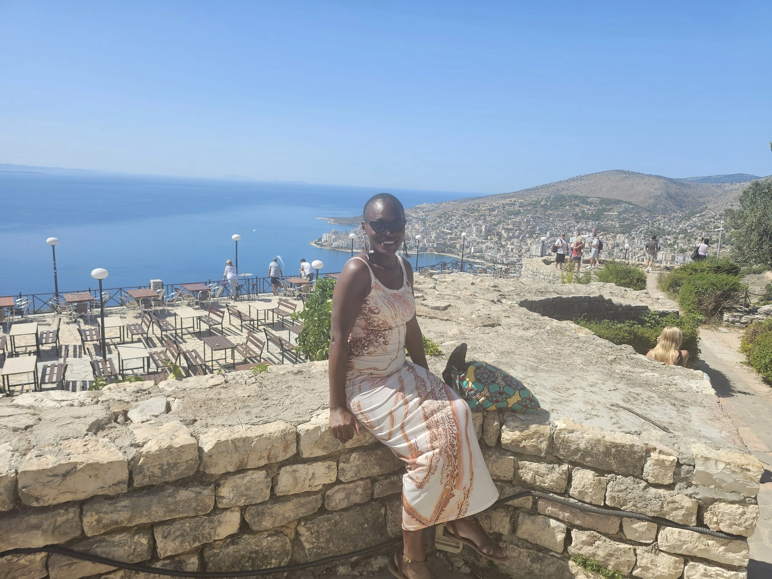 Femme assise sur un mur en pierre avec vue sur la mer et la ville, en vacances, portant une robe d'été, lunettes de soleil, sac coloré, journée ensoleillée.
