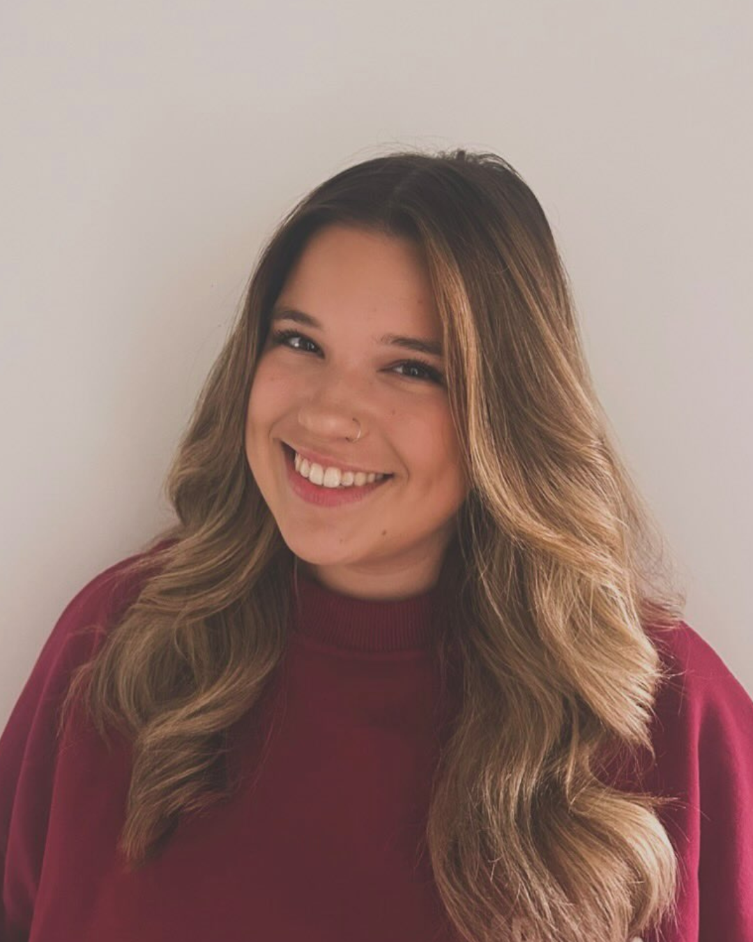 Caucasian girl with blonde brown long hair smiling at the camera wearing a red jumper in front of a beige background