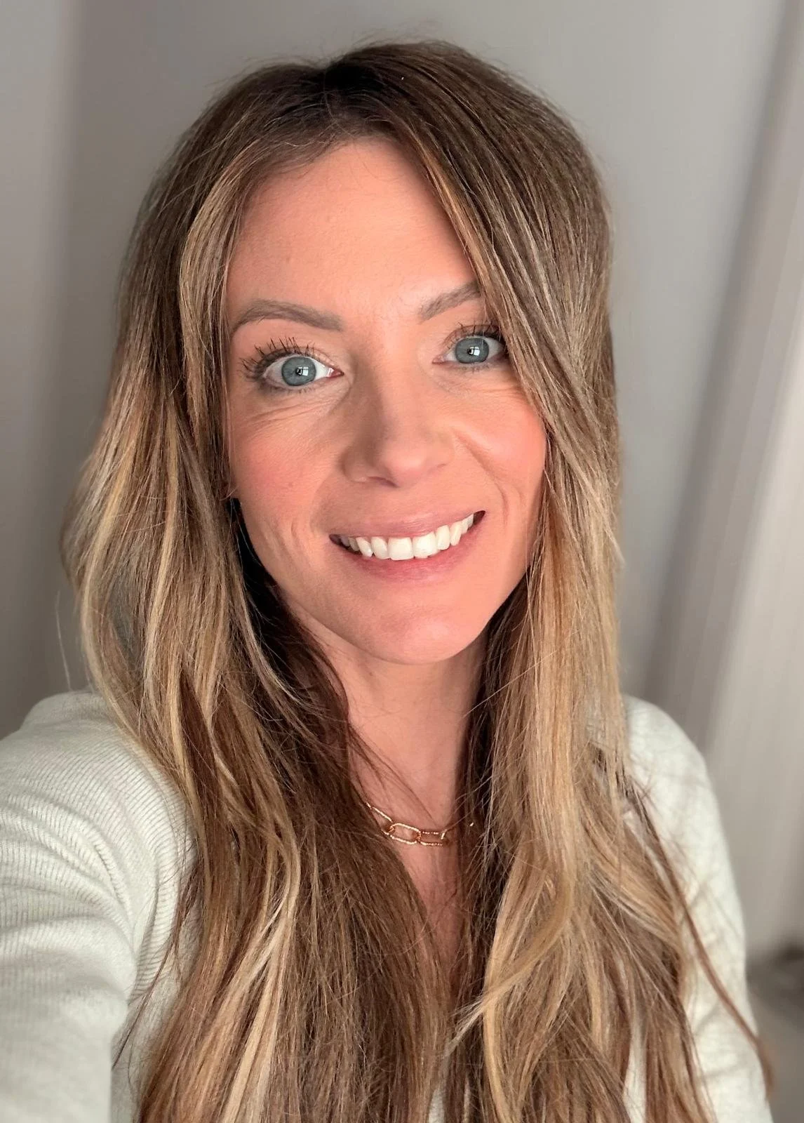 A woman with long, wavy brown hair and blue eyes smiling at the camera. She is wearing a white top and a gold necklace. The background shows a plain wall and part of a doorframe. This is Jody.