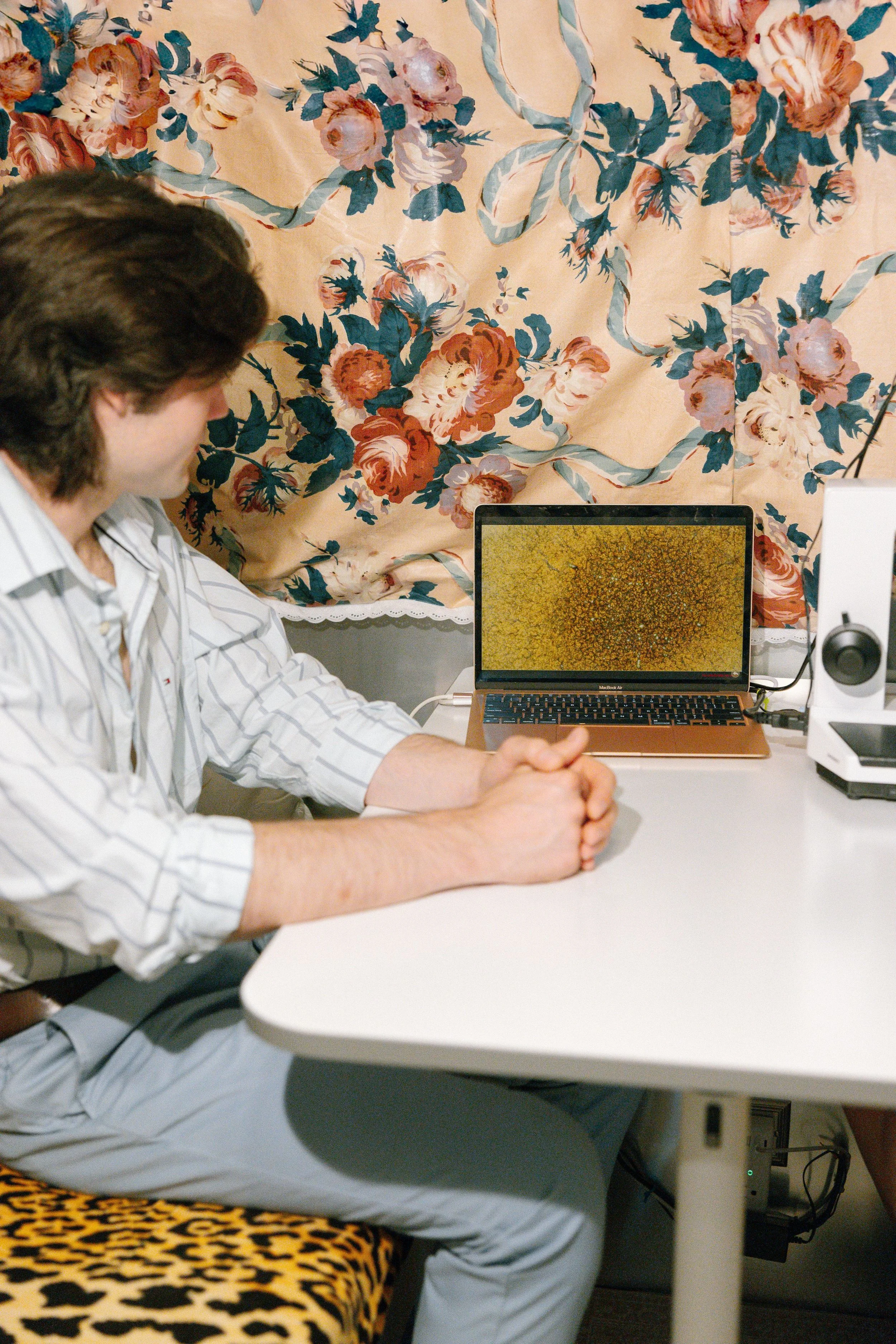 A man is sitting at a desk in front of a laptop with a colorful abstract wallpaper on the screen. He is wearing a white striped shirt and gray pants, with his hands clasped on the desk. Behind him, there is a floral curtain with a large pattern of pink and white flowers with green leaves on a beige background.