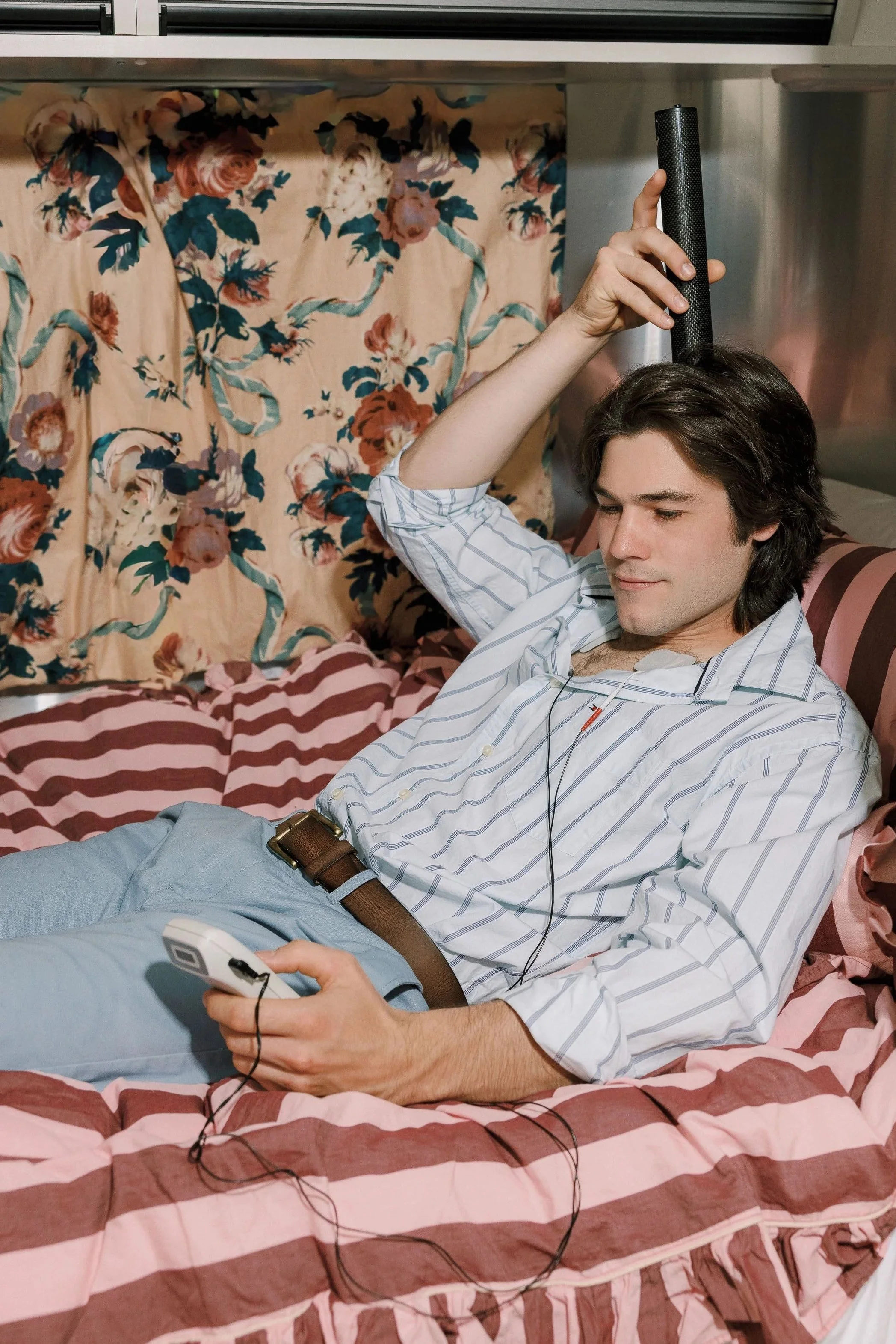 A young man with dark hair, wearing a striped shirt, is lying in bed while holding a TV remote in his right hand and a smartphone connected to earphones in his left hand. He appears relaxed, resting on a bed with a pink and brown striped blanket and floral curtains in the background.