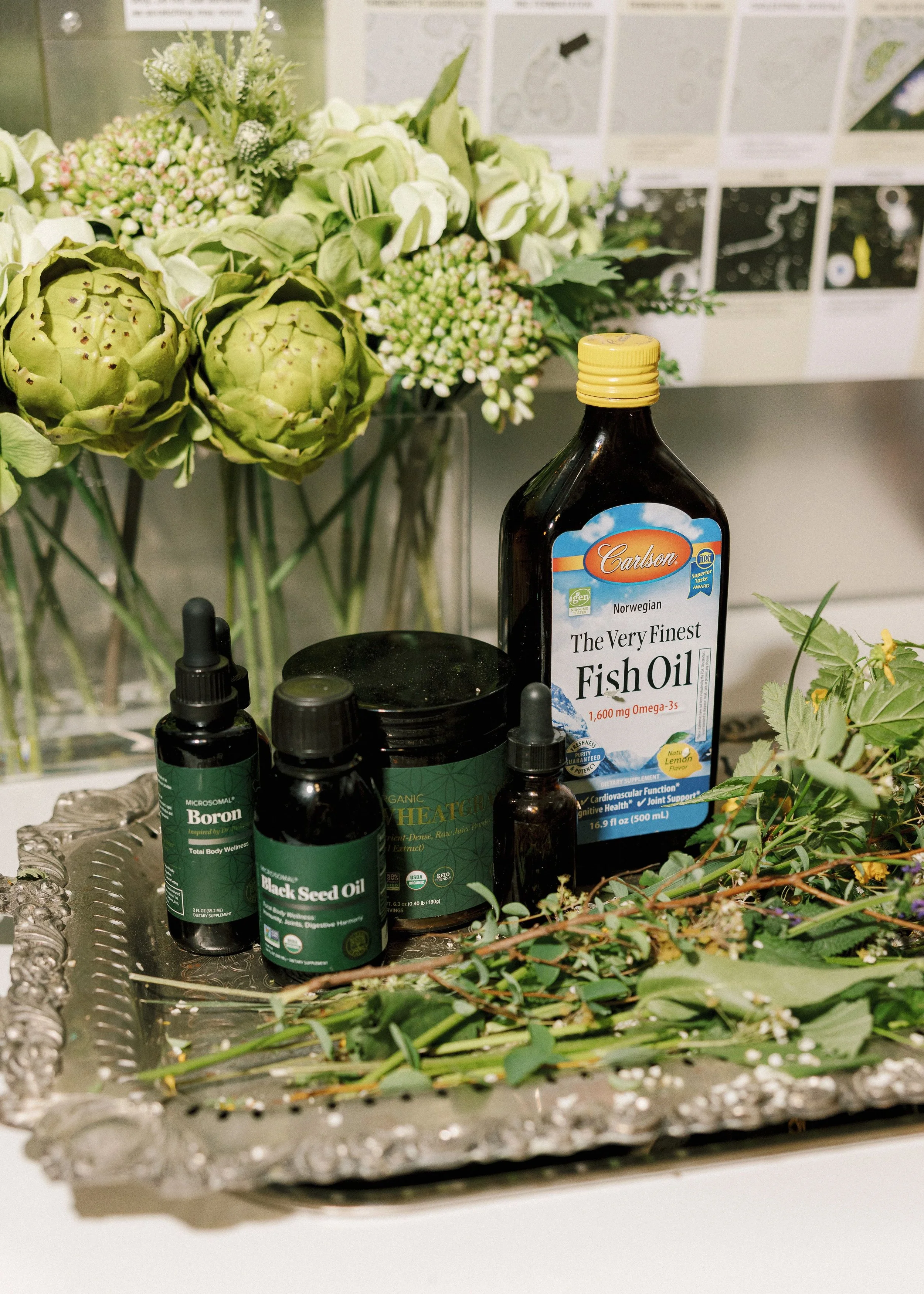 A floral arrangement consisting of artichokes, white roses, and small white and pink flowers in a vase. In the foreground, there are bottles of supplements including fish oil, black seed oil, and boron, along with some botanical elements and a decorative silver tray.