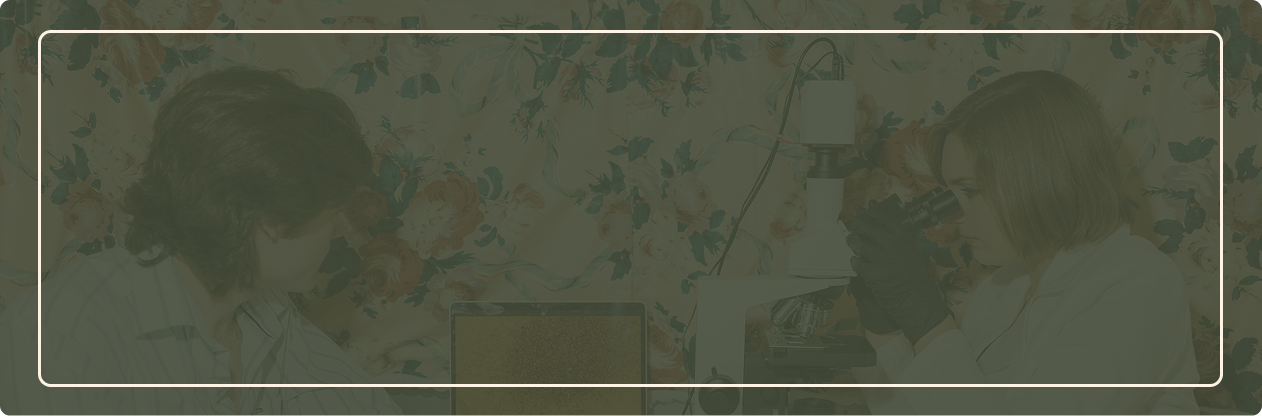 A scientist and a researcher wearing gloves look into a microscope in a lab with floral wallpaper.