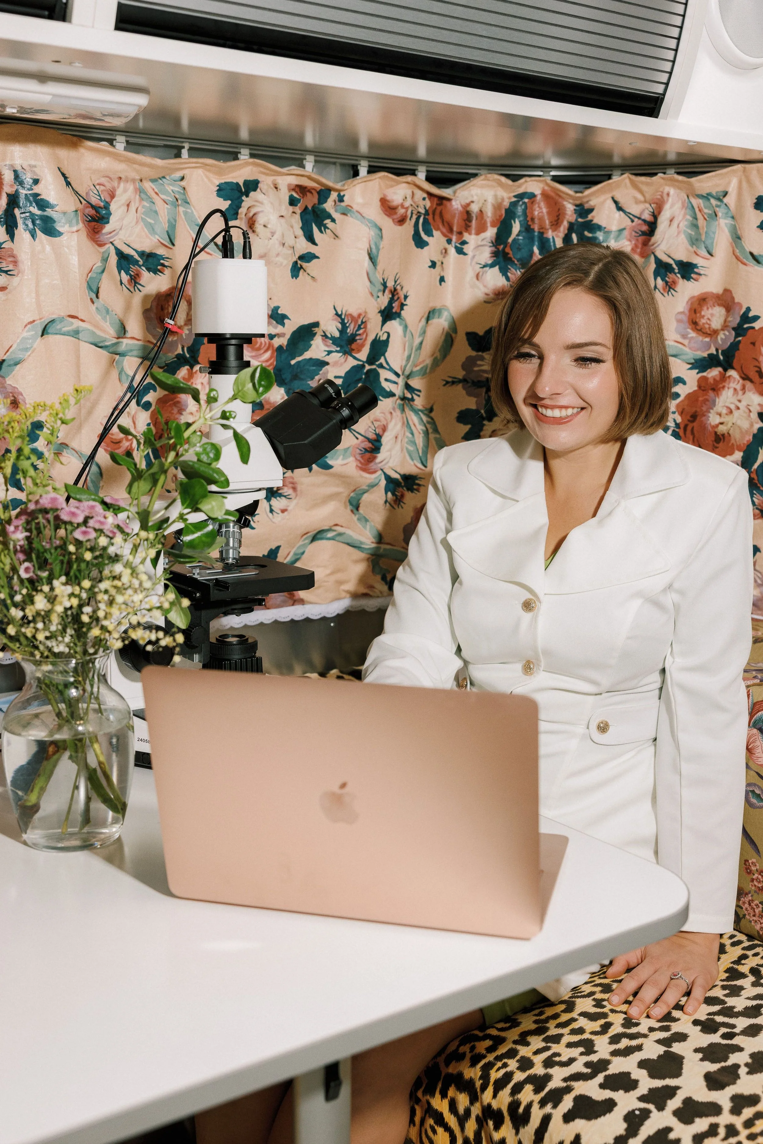 A woman smiling as she sits at a white table with a pink laptop in front of her. There are colorful flowers in a glass vase on the table. Behind her, there is a floral-patterned fabric backdrop, a microscope, and some electronic equipment.