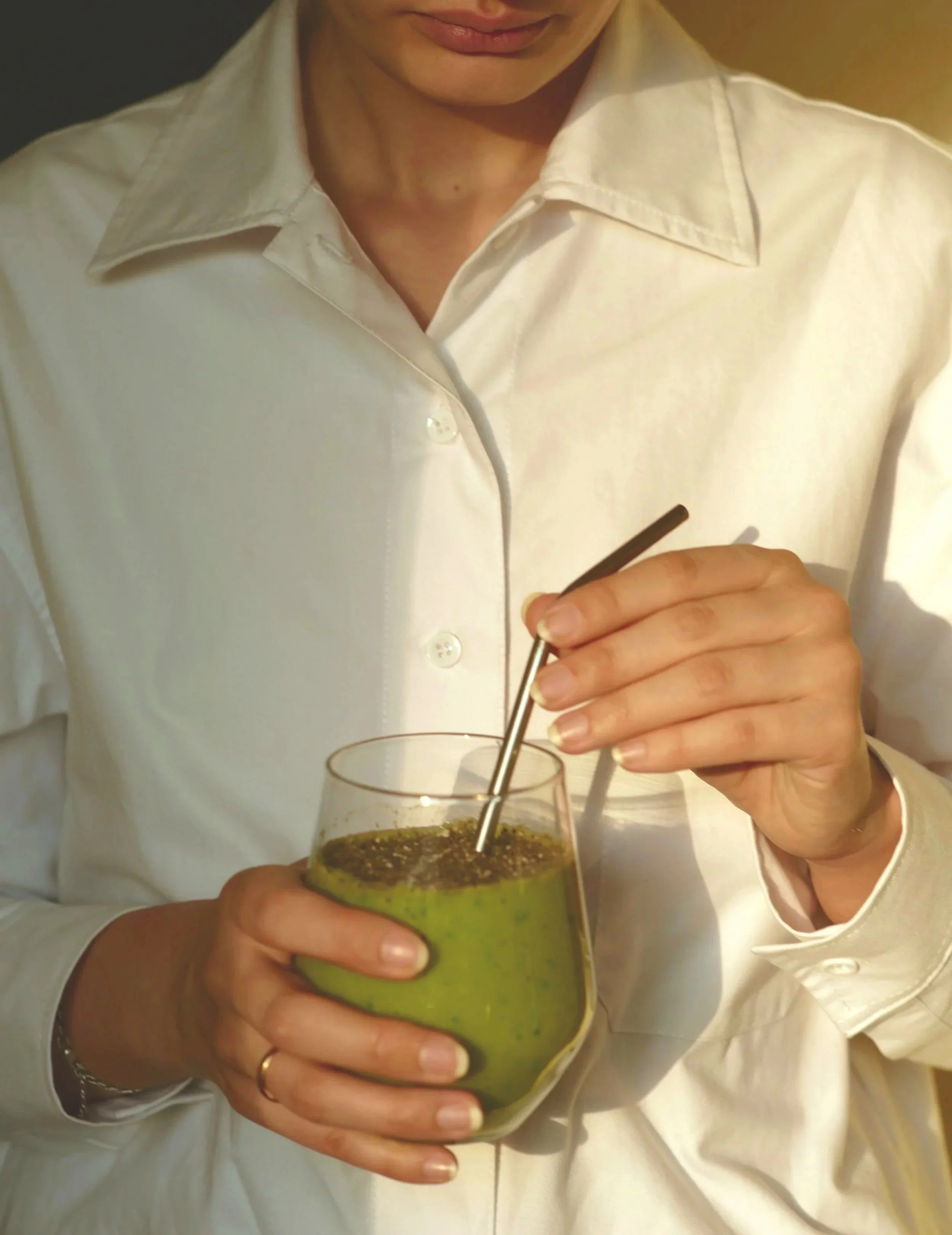 Close-up of a woman in a white shirt holding a glass of green juice, with a silver straw, while stirring it with a black stick or straw.
