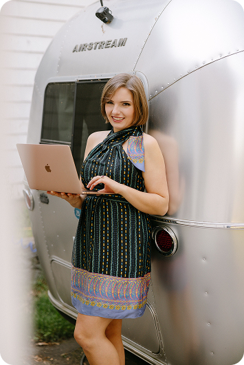 Woman standing outside next to an Airstream trailer, holding a laptop, smiling at the camera.
