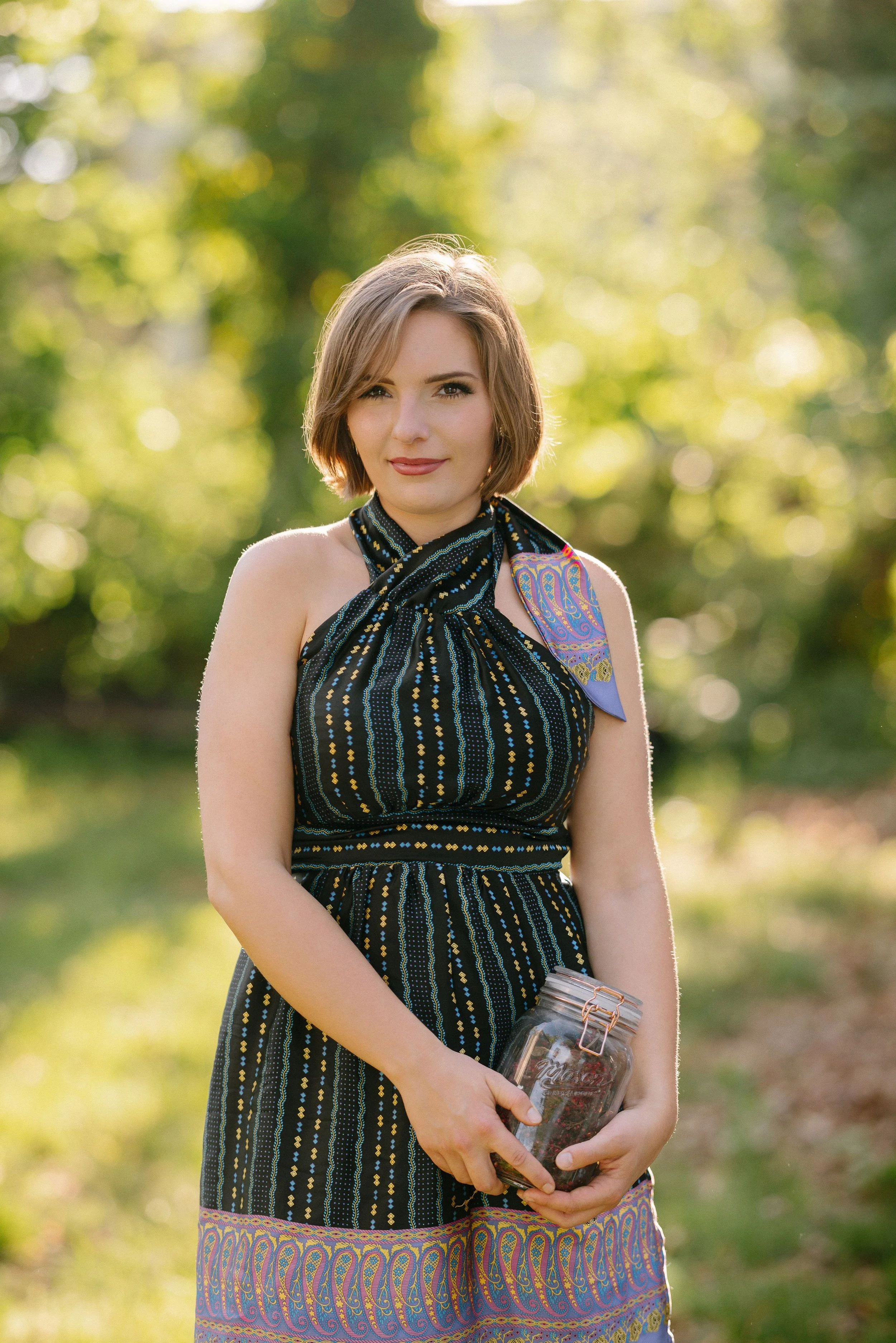 A young woman with short brown hair and light skin, wearing a dark patterned dress with shoulder straps and a colorful scarf, stands outdoors holding a glass jar with a metal lid and straw, with a blurred green park background and dappled sunlight.