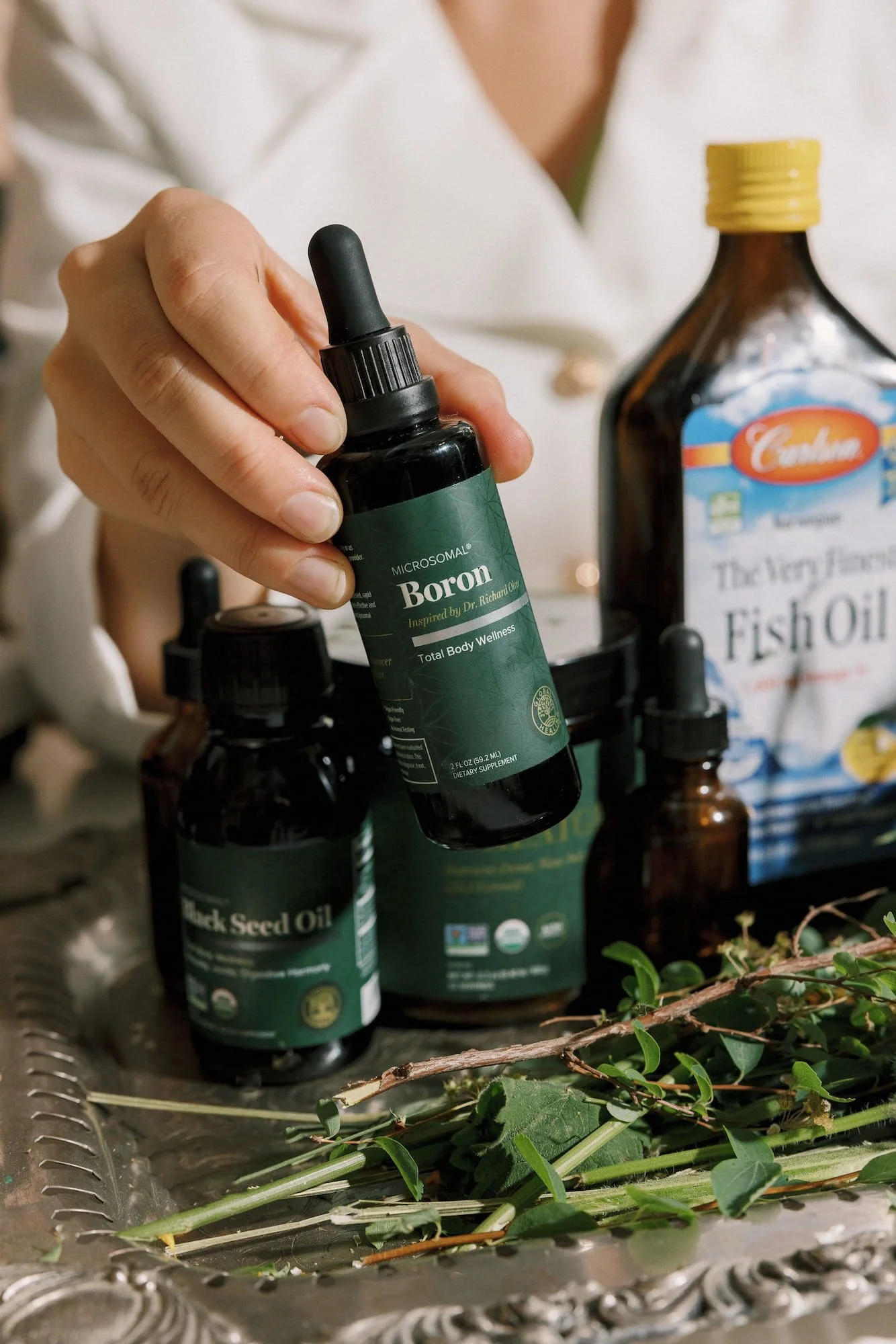 A person holding a small dark bottle labeled 'Boron,' with other bottles and a bag of fish oil in the background, on a silver tray with green plant cuttings and twigs.