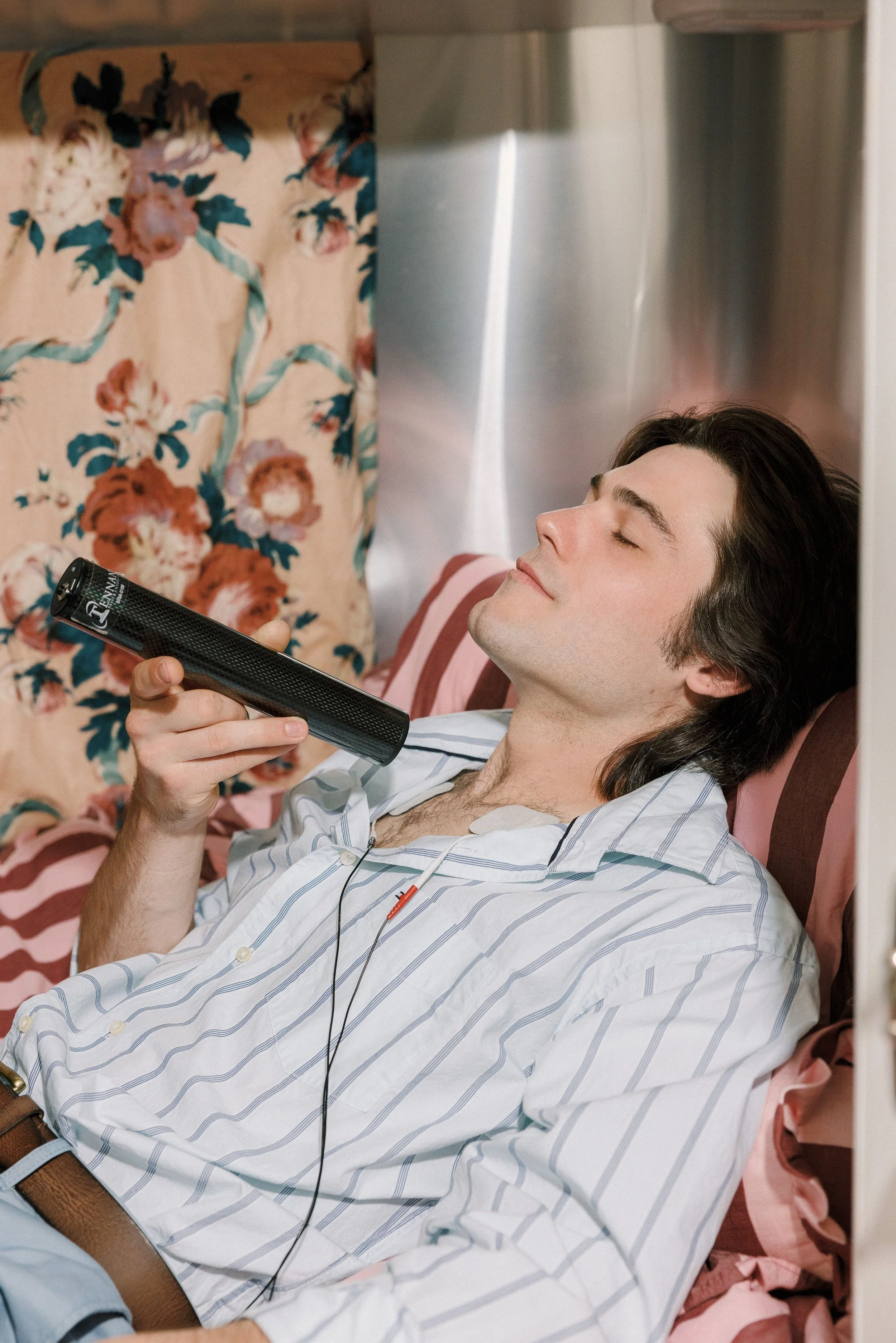 A young man with long dark hair is lying in a hospital bed, resting, with his eyes closed. He is holding a black flashlight in his right hand, and wearing a striped hospital gown. There are floral-patterned curtains near the bed, and a silver metallic wall or panel behind him.