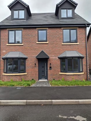A three-story brick house with black window frames and a black front door, two dormer windows on the roof, and small bay windows on the ground floor. There is a sidewalk and grass in front.