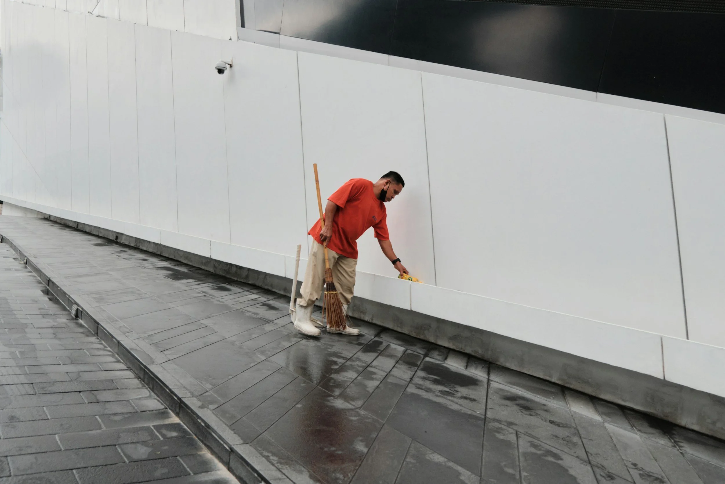 A man in a red shirt and beige pants sweeping a wet tiled sidewalk next to a white wall, with a broom and a dustpan.