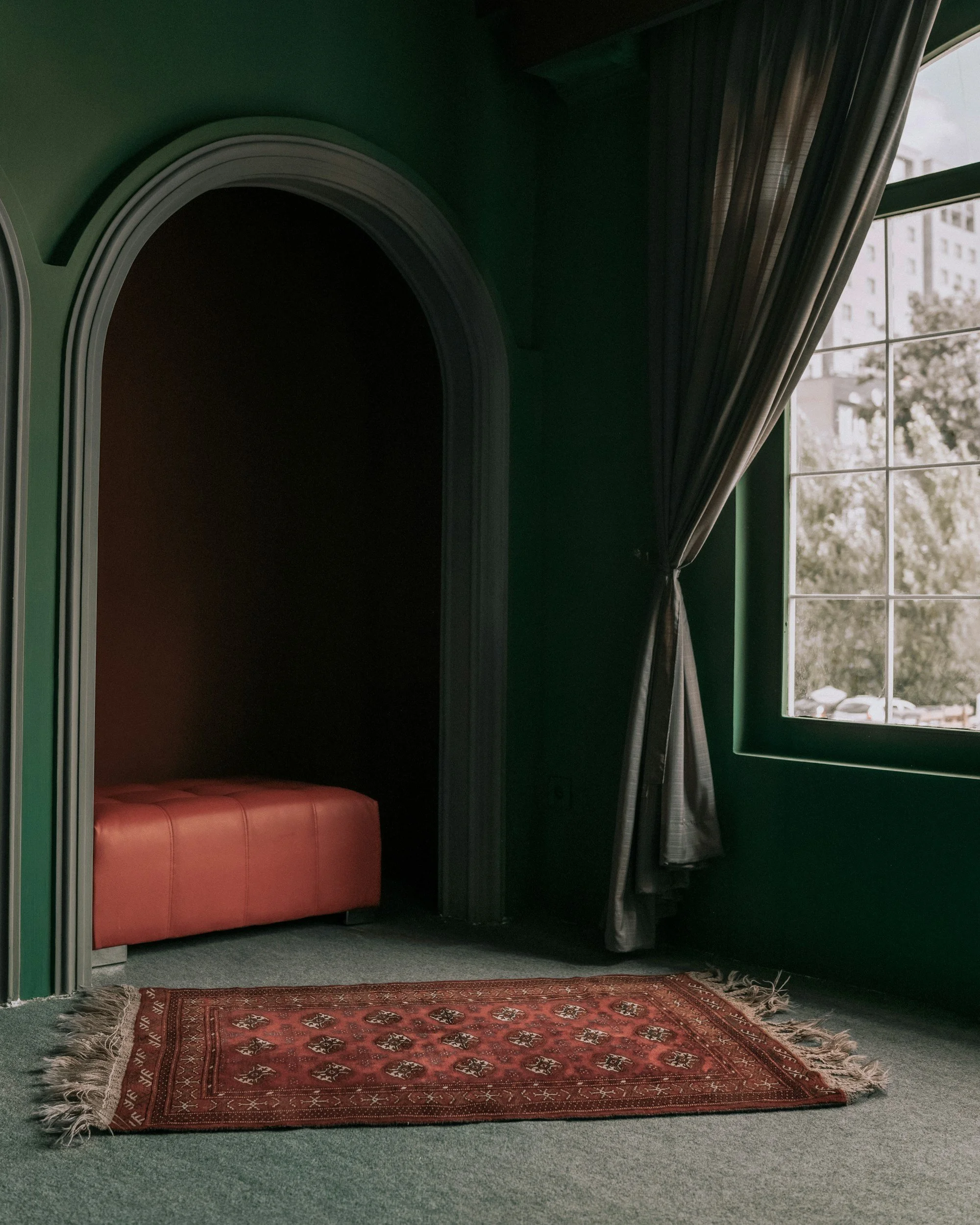 A room with dark green walls, a red ottoman, a patterned red rug on gray carpet, large window with black curtains, and a view of buildings and trees outside.