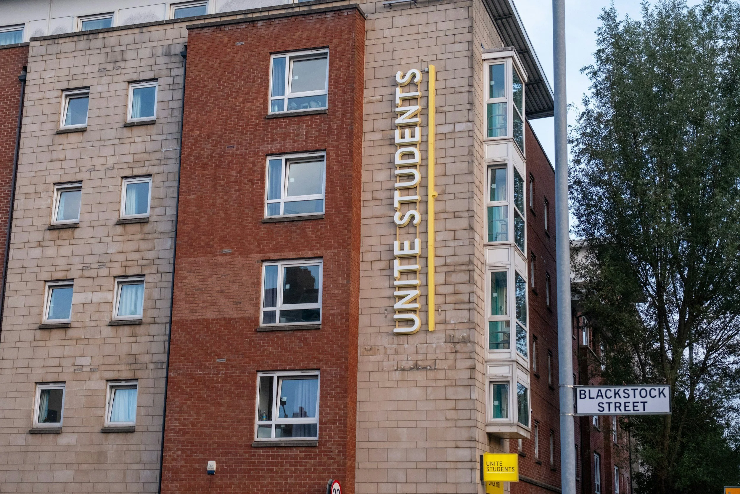 A city building with the sign 'UNITE STUDENTS' on the corner, and a street sign reading 'BLACKSTOCK STREET.' The building has a mixed facade of beige and red bricks with windows, some with blue curtains and a corner bay window.