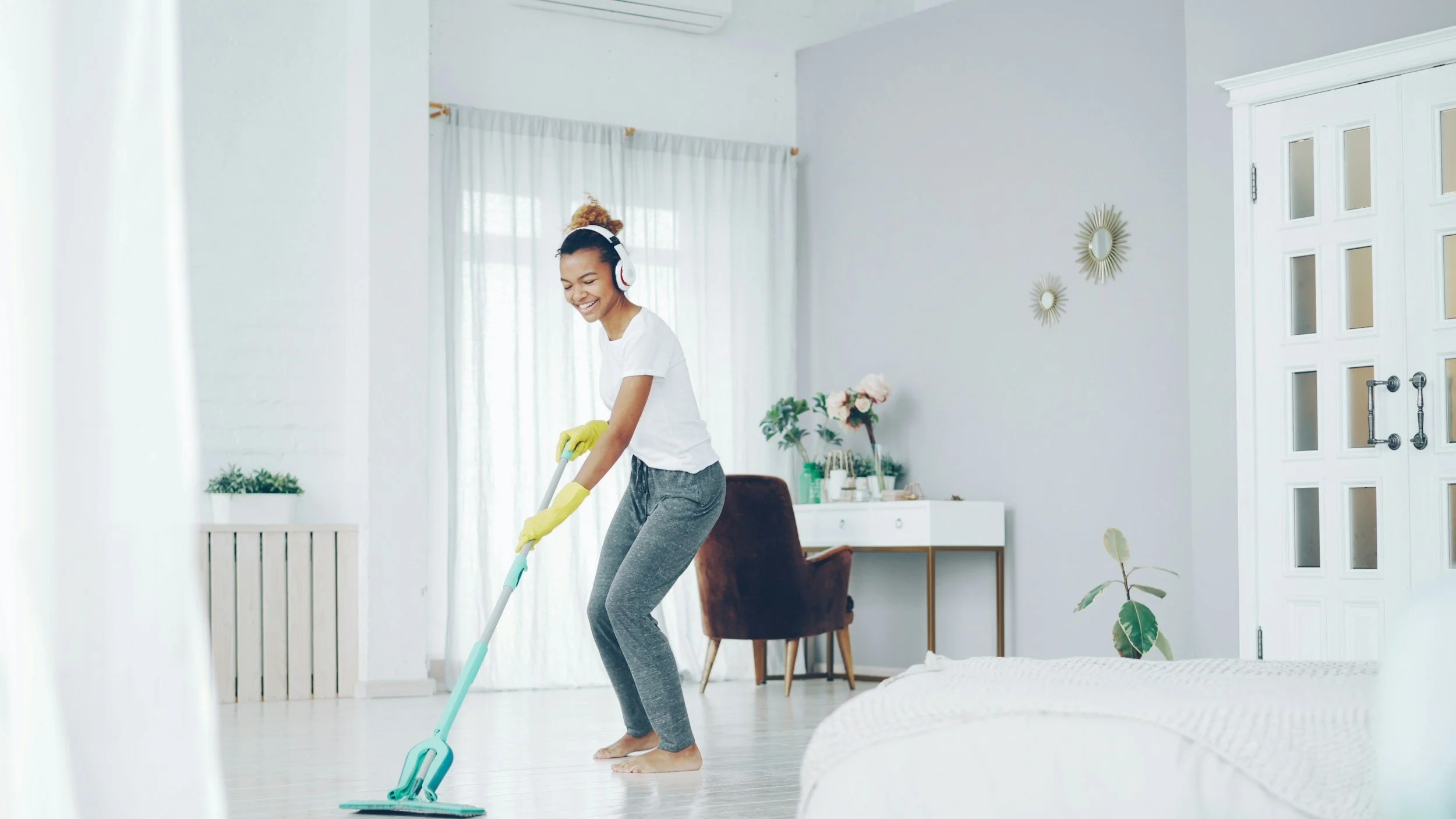 Woman smiling while vacuuming the floor in bright, modern living room, wearing headphones and yellow gloves.