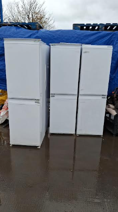 Three white refrigerators standing outdoors on a wet ground with a blue tarp and trees in the background.