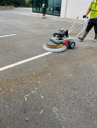 A street cleaner is using a ride-on sweeping machine to clean the parking lot near a building, with a person walking in the background.