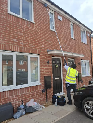 Worker in a high-visibility vest cleaning the exterior windows of a brick residential building using a long extension pole, with tools and equipment on the ground nearby.