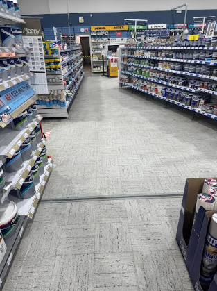 A supermarket aisle with shelves stocked with bottled products, travel-sized items, and other groceries, with a shopping cart and checkout counter in the background.