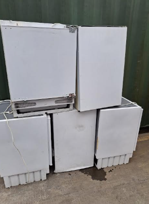 Four white mini refrigerators placed on the ground outdoors, with a green fence in the background.