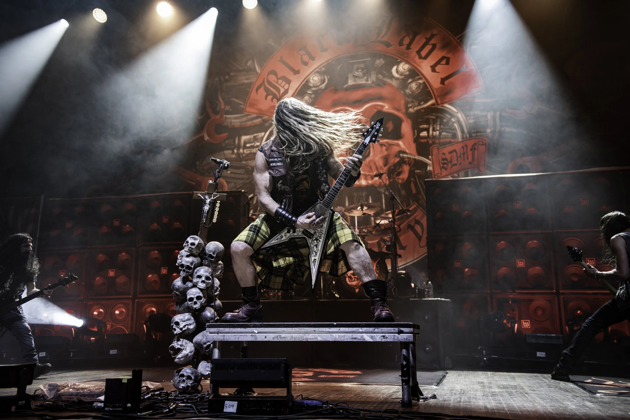 A rock band performing on stage with dramatic lighting and skull decorations. A guitarist in plaid shorts and long hair is at the center.