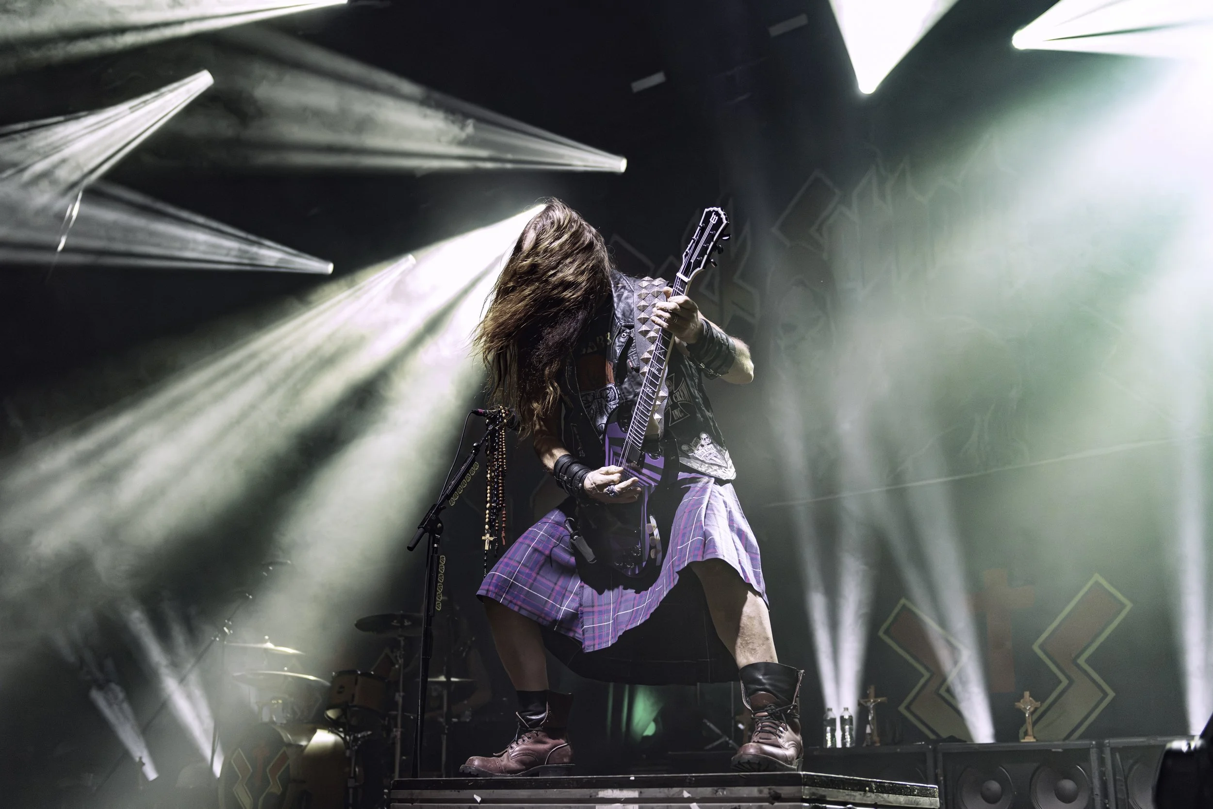 A musician with long hair playing an electric guitar on stage, with stage lights and smoke effects.