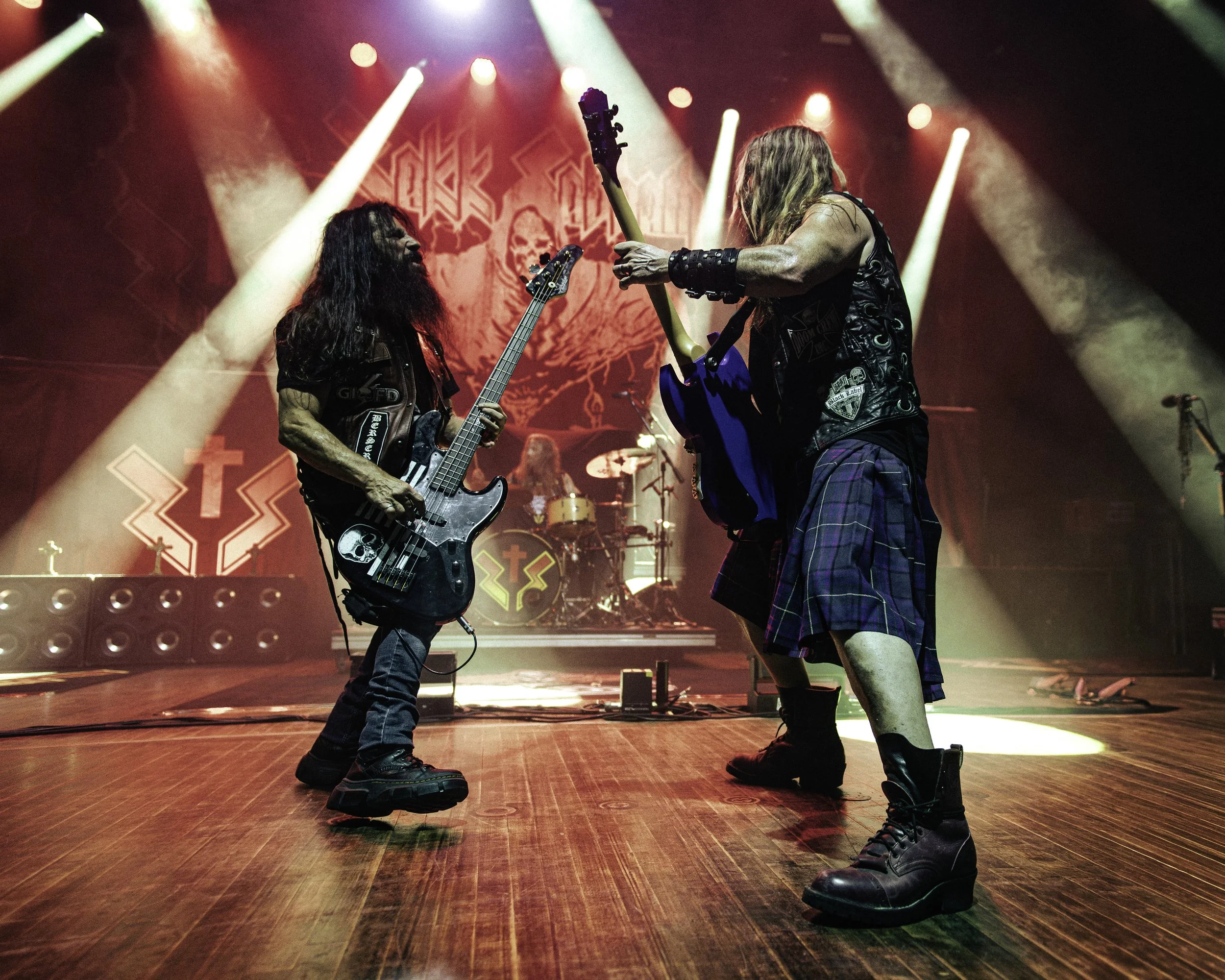 Two male musicians performing on stage with guitars, wearing punk-rock style leather vests and boots. The stage is illuminated with colorful lights, and the background features a large skull logo with red and black designs.