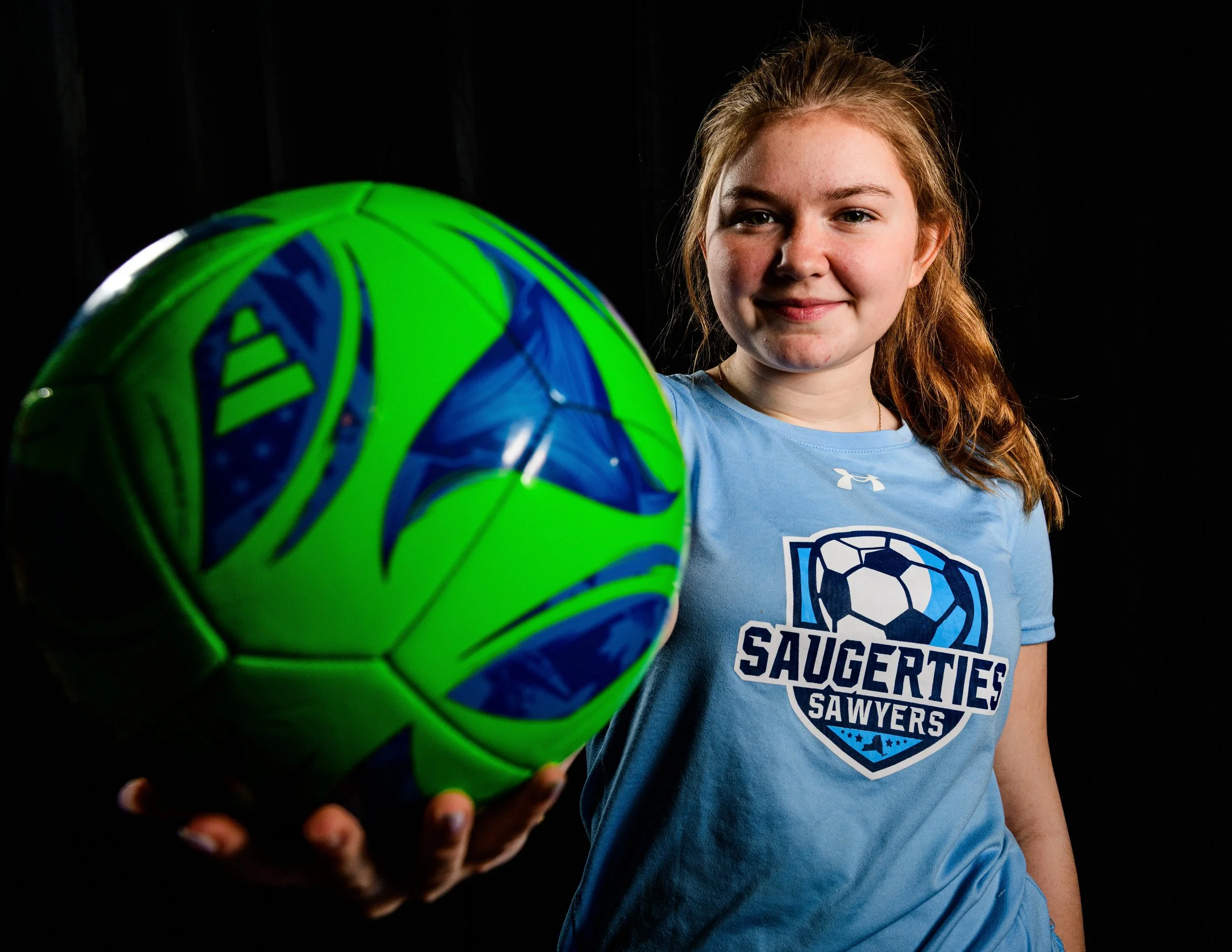 A young girl with red hair smiling and holding a green and blue soccer ball towards the camera, wearing a light blue soccer jersey with a soccer ball emblem and the text 'Saugerties Sawyers' on it, against a dark background.