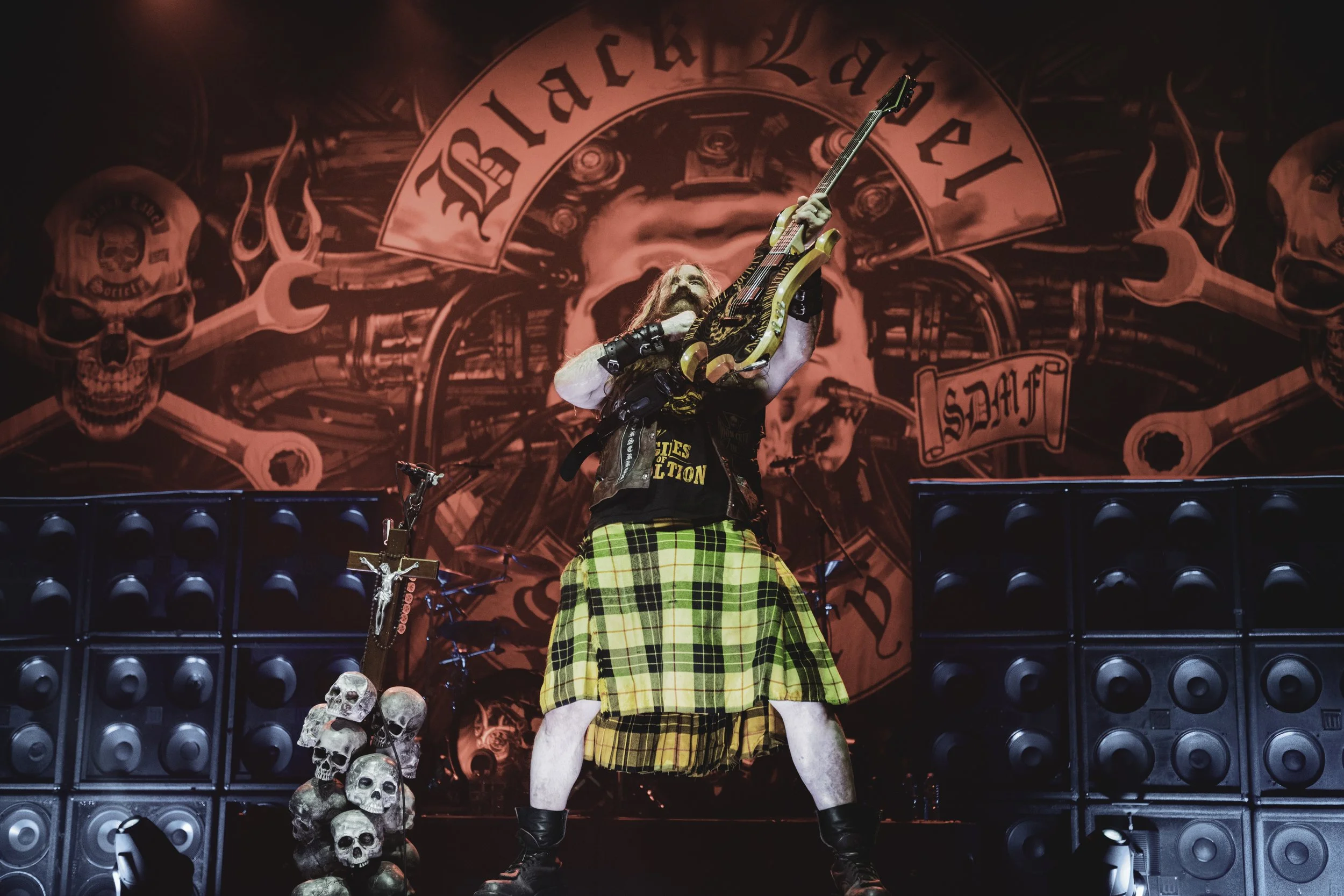 A man with long hair and beard holding an electric guitar pointing it upward. He is wearing a black t-shirt with a graphic design, a kilt with a tartan pattern, and black boots. The stage has skull prop decorations on the floor and large speaker stacks. The background features a red and black banner with skulls and fire graphics.