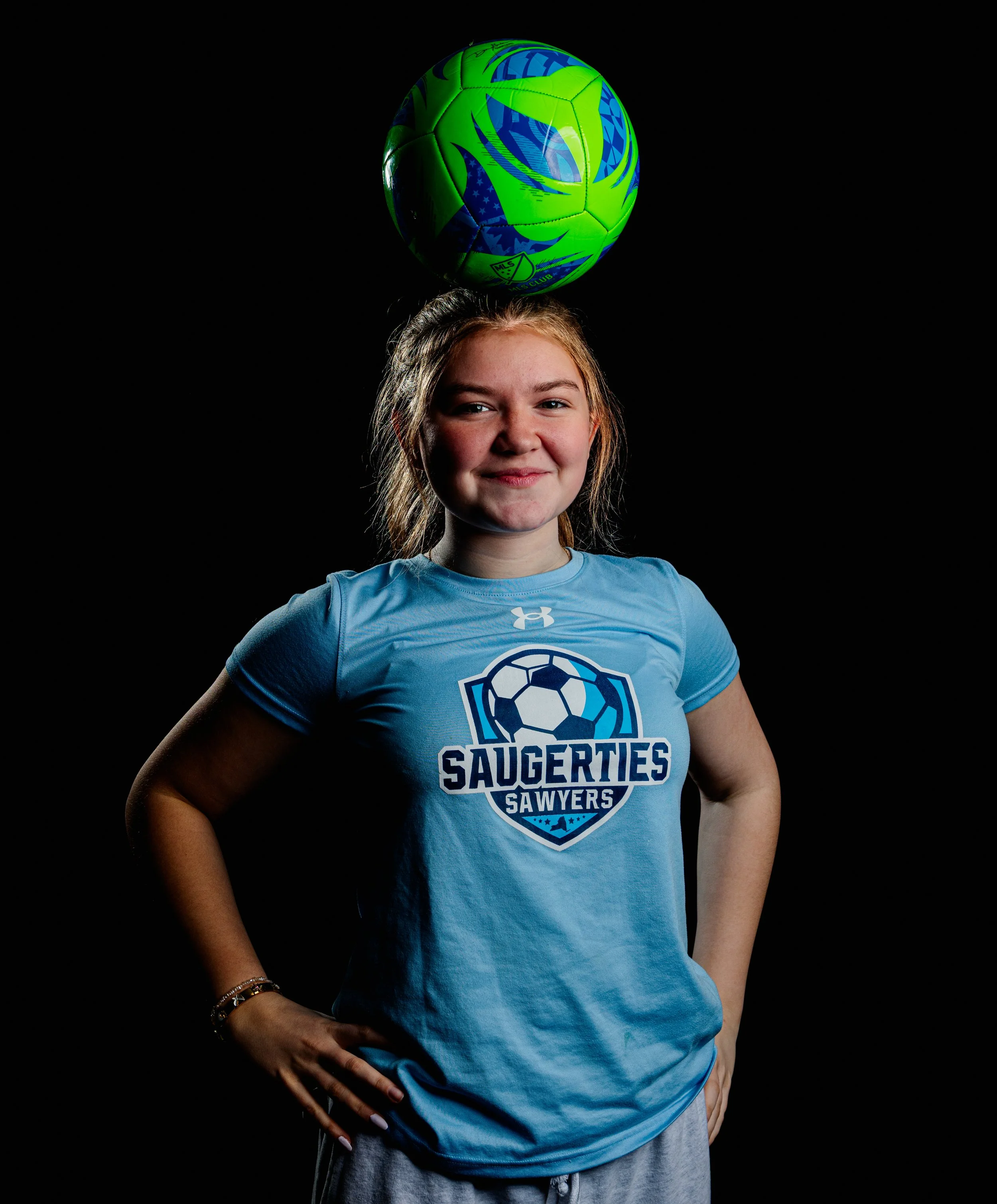 Young girl smiling, wearing a blue sports shirt with 'Saugerties Sawyers' logo, balancing a green and blue soccer ball on her head, against a black background.