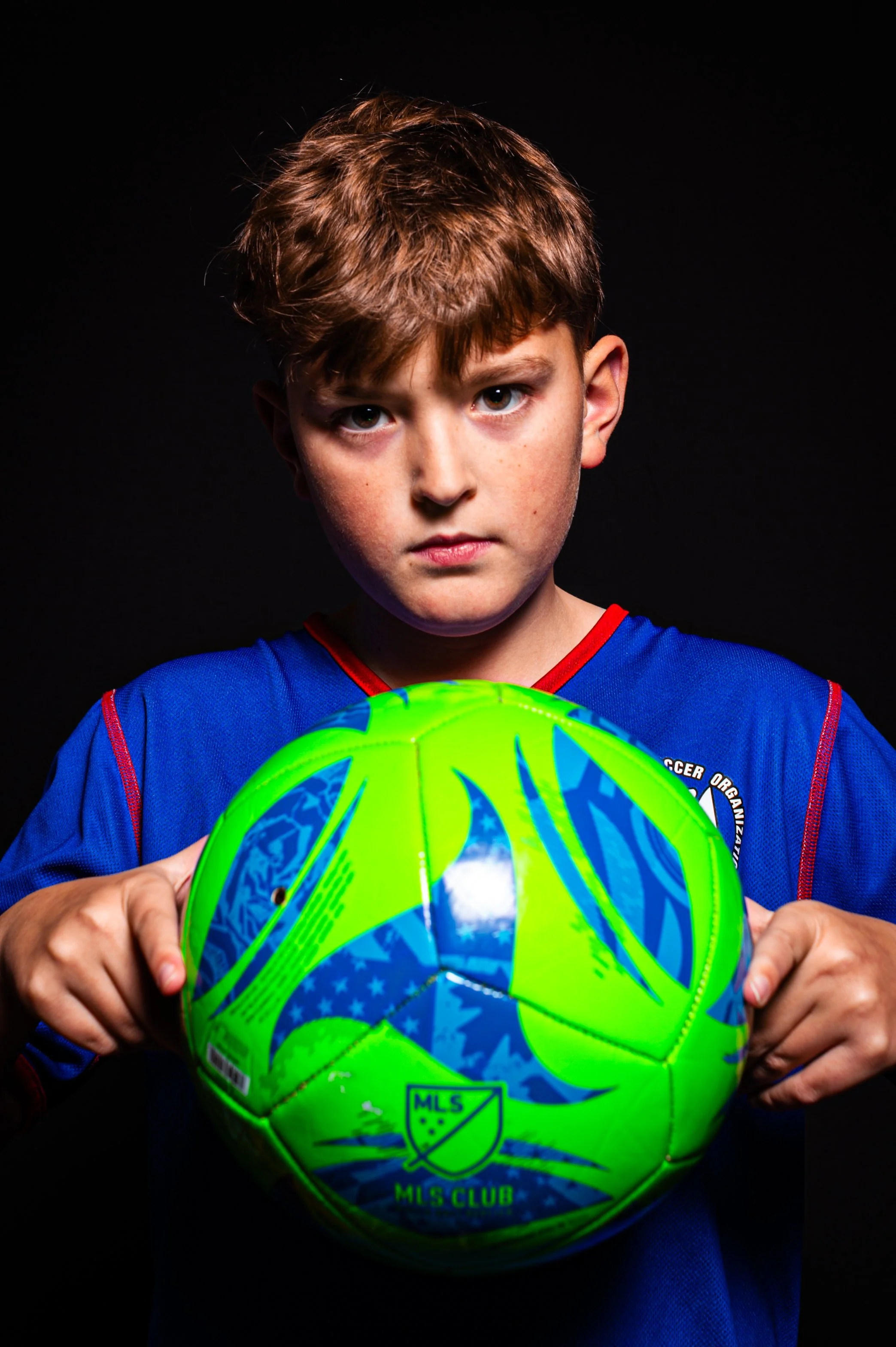 A young boy wearing a blue sports jersey holding a colorful green and blue soccer ball with the MLS logo on it, against a black background.
