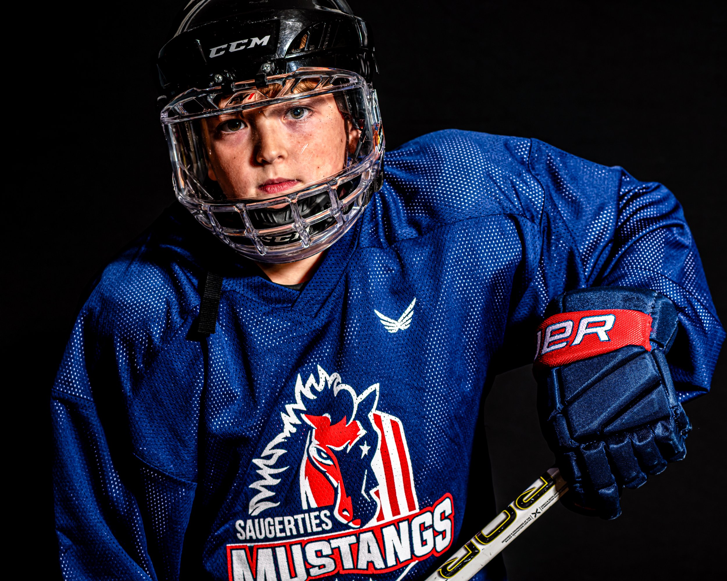 Young hockey player in a blue jersey with a mustang logo and 'Saugerties Mustangs' text, wearing a black helmet and protective gear, holding a hockey stick.