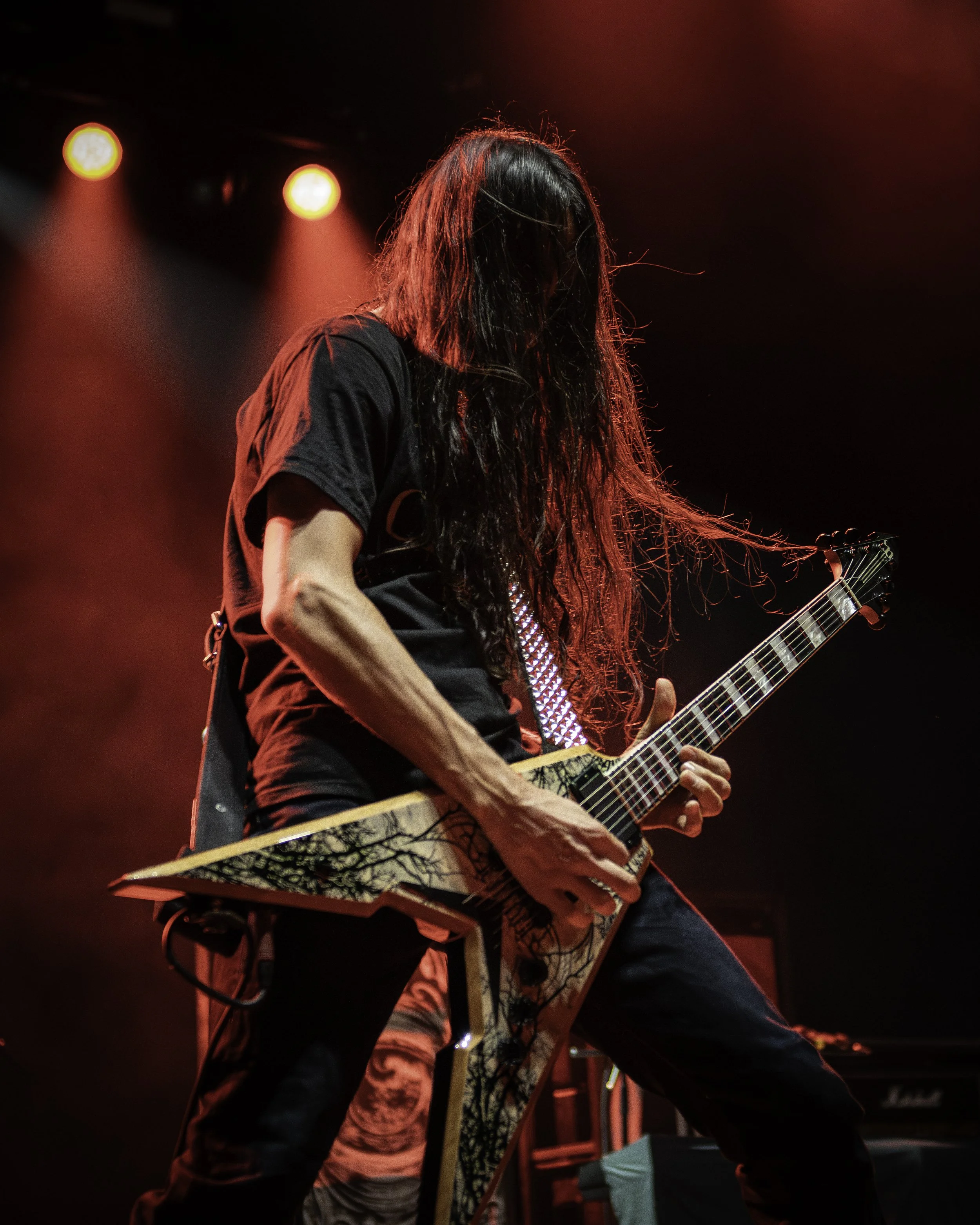 A musician with long hair playing an electric guitar on stage, illuminated by red stage lighting.