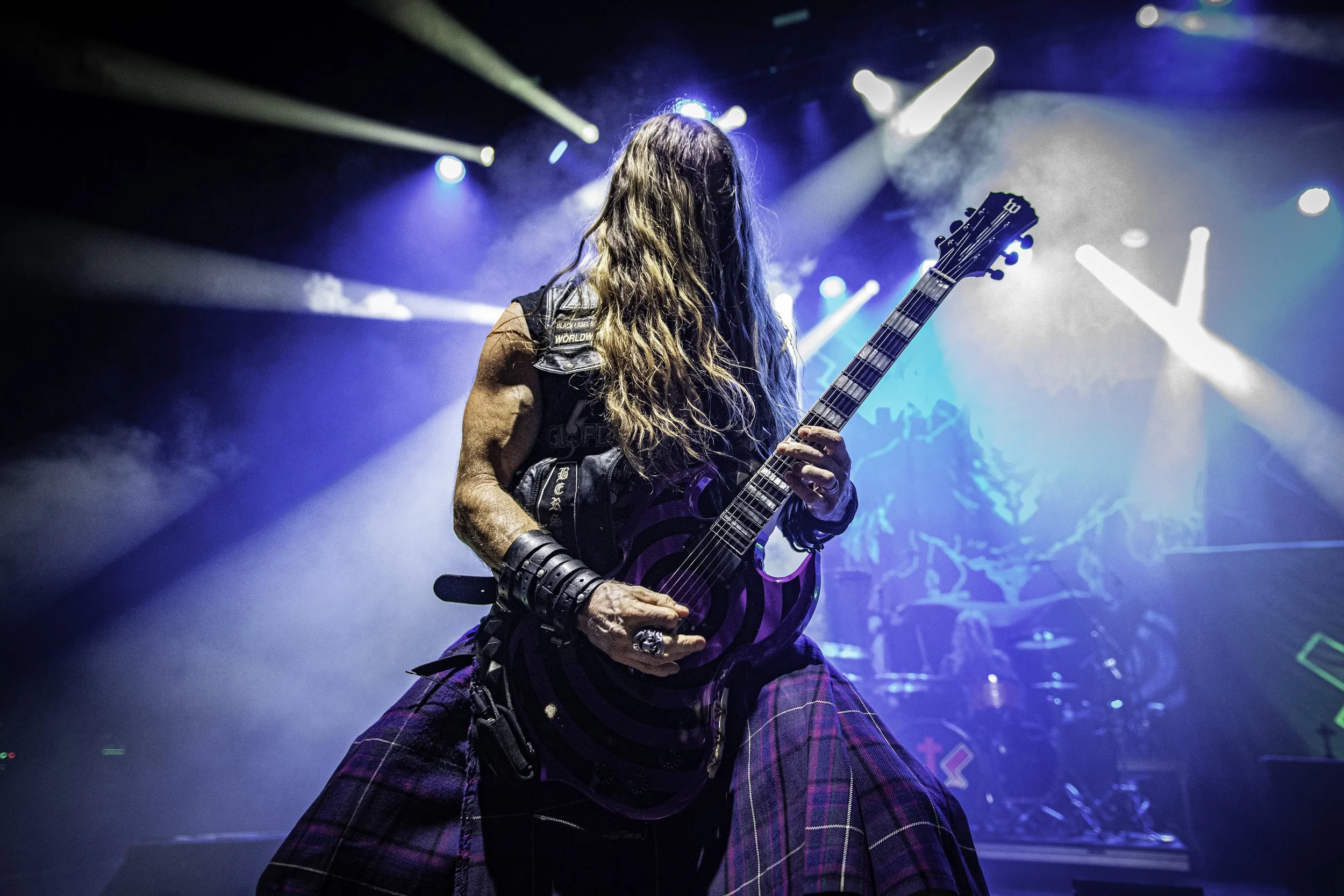 A long-haired musician playing an electric guitar on stage with colorful lights and smoke effects.