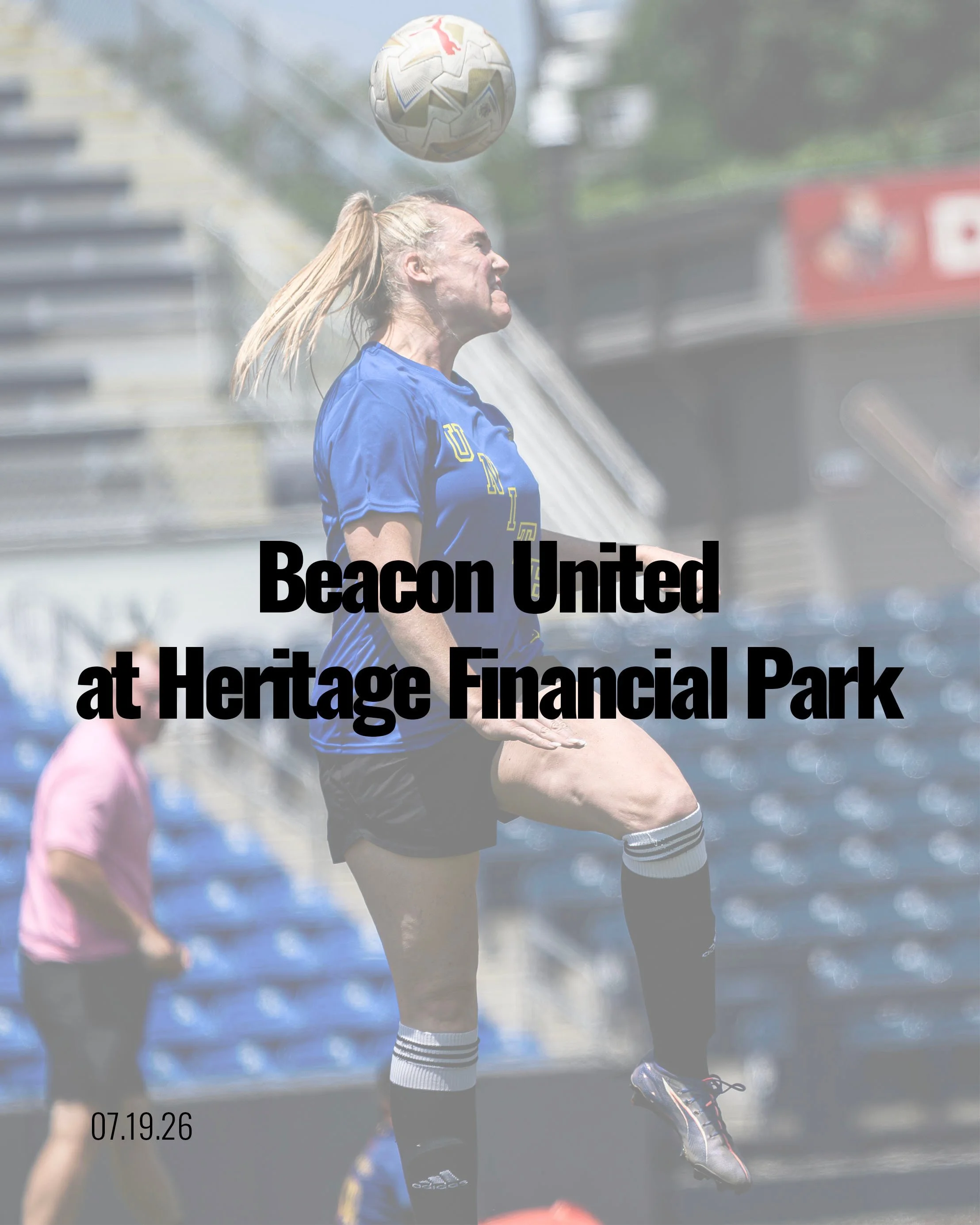 A female soccer player in a blue jersey and black shorts is mid-air, heading a soccer ball during a game outdoors on a bright day. A person in a pink shirt can be seen in the background, and the scene appears to be at Heritage Financial Park.