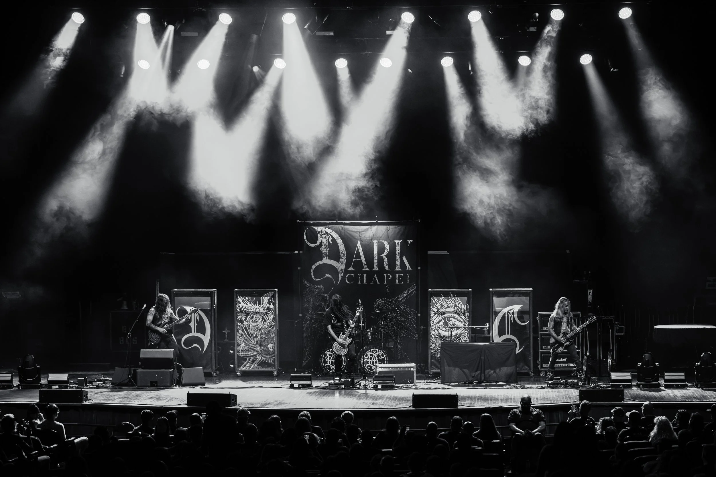 A band performing on stage at Dark Chapel with musical equipment and decorated backdrop, illuminated by stage lights, with audience seated in front.