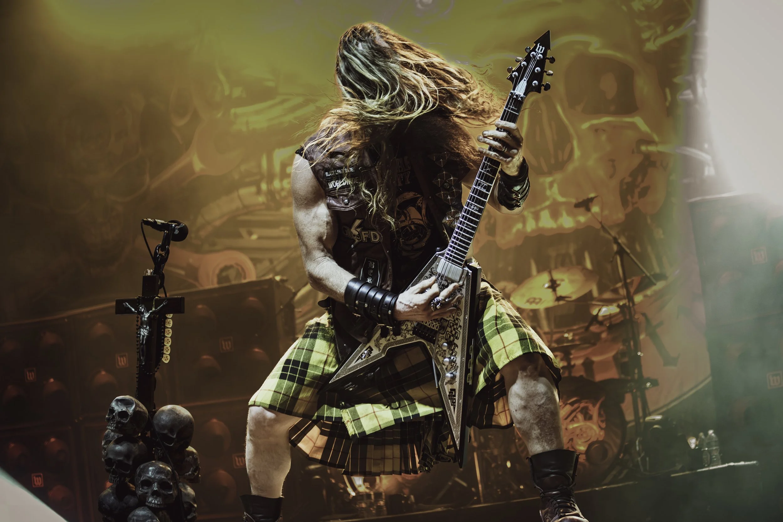 A guitarist performing on stage, wearing a black vest with patches and yellow tartan shorts, with long hair covering his face, in a concert setting with drums and skulls on the floor, and a fiery background.