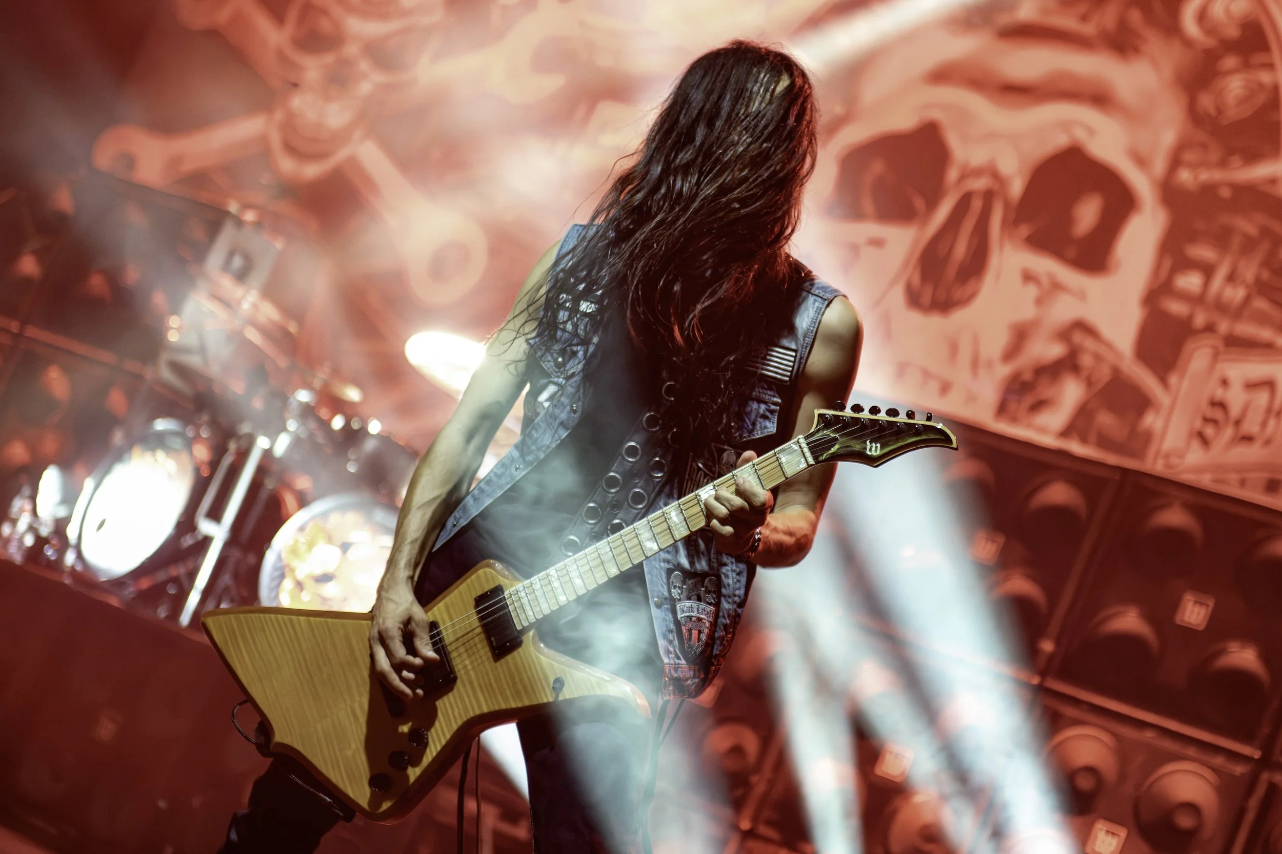 A female guitarist performing on stage with long dark hair, wearing a sleeveless denim vest, and playing an electric guitar. The background shows red lights and abstract visuals.