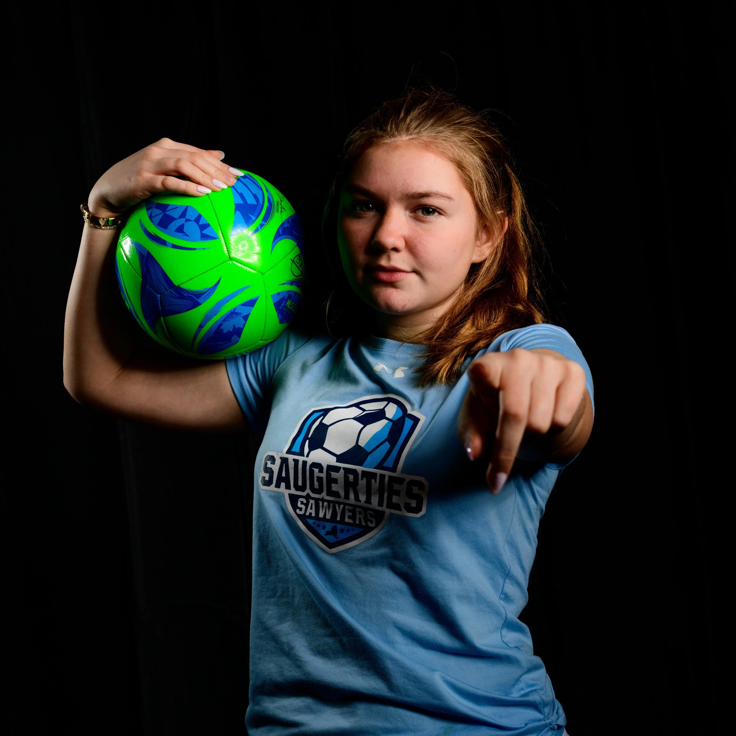 A young female soccer player in a blue jersey pointing forward while holding a green and blue soccer ball on her shoulder against a black background.