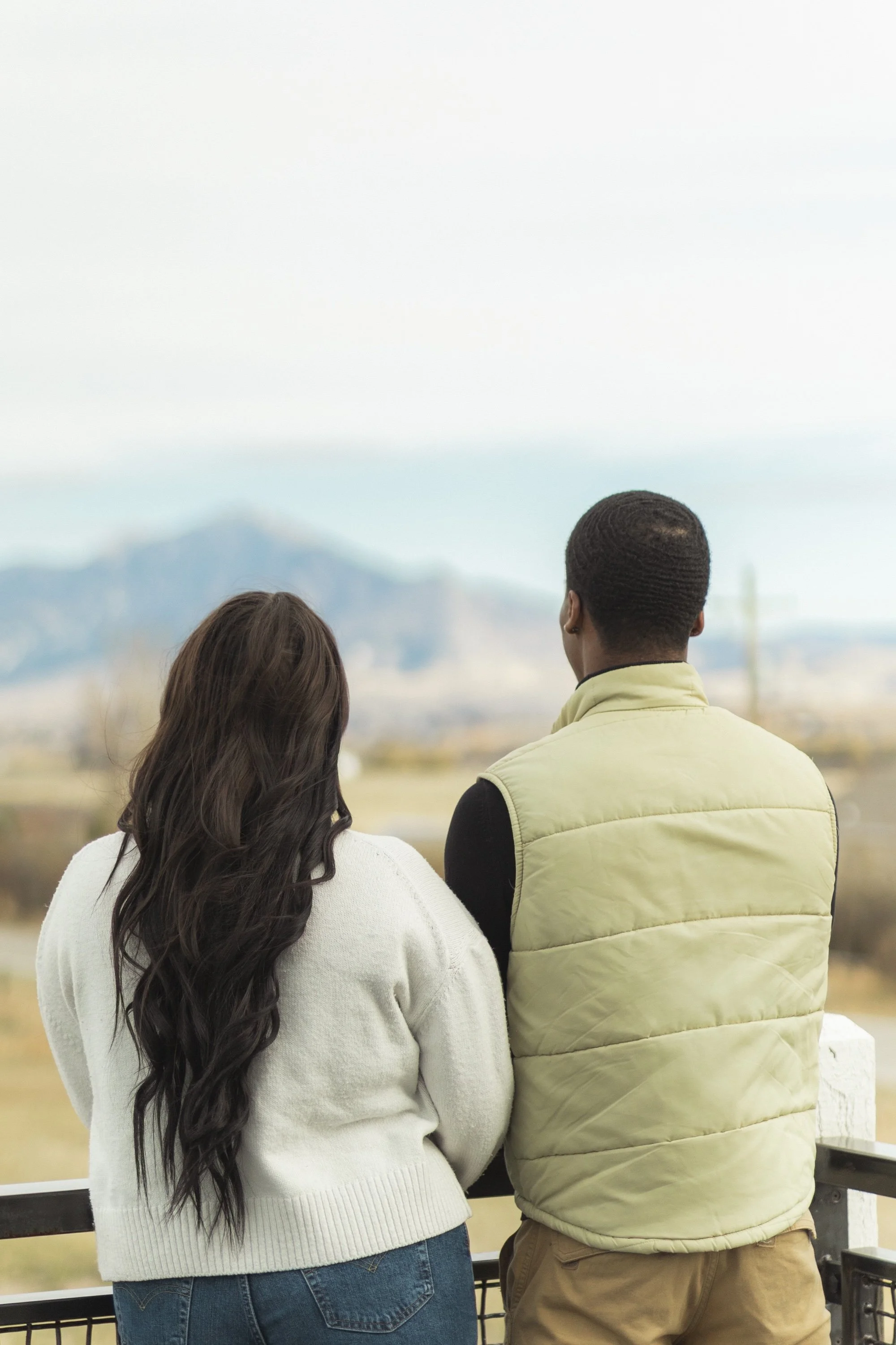 Two people standing on a balcony, looking at mountain scenery in the distance.