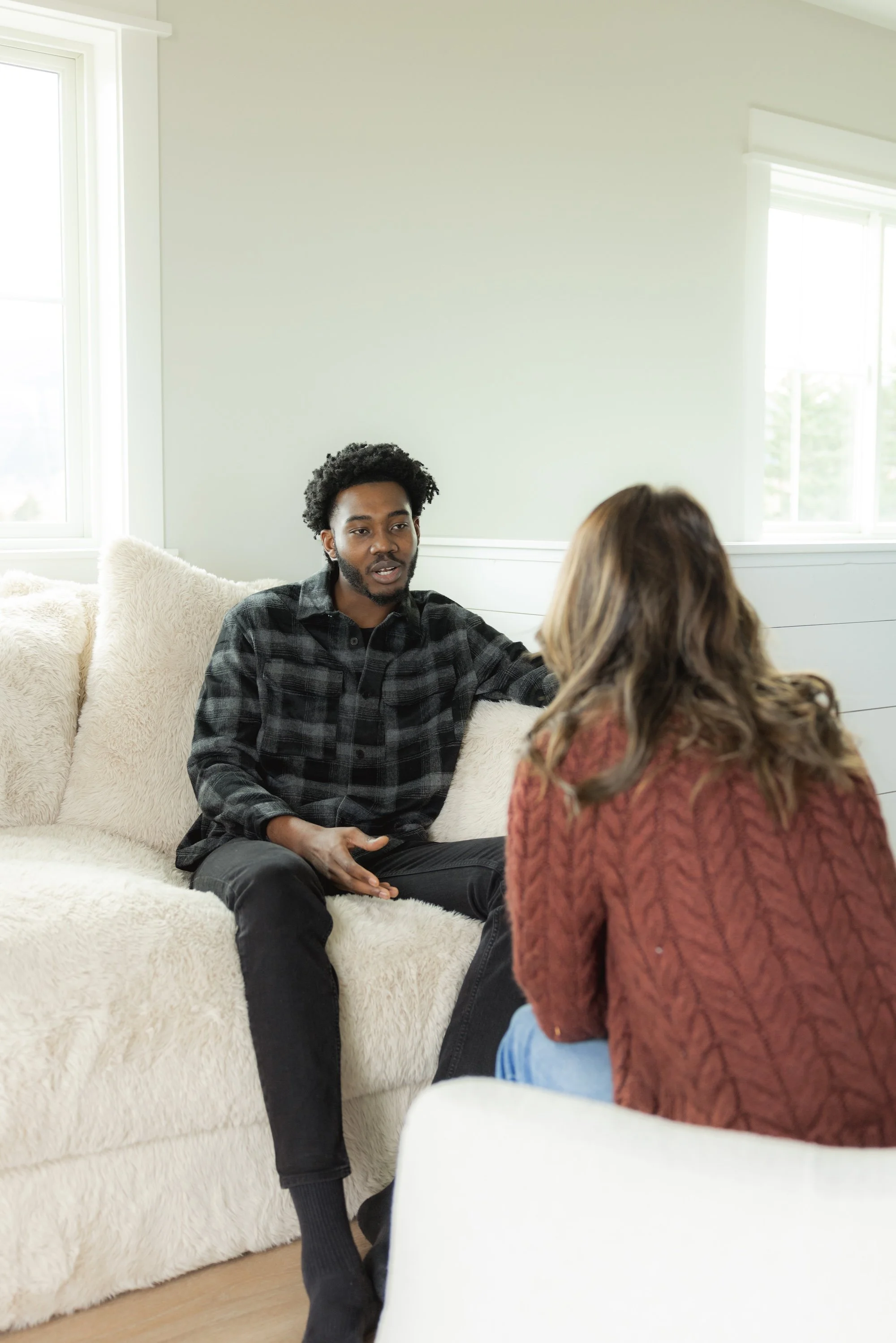 Two people having a conversation in a living room with natural light from windows, one sitting on a cream-colored couch and the other facing away from the camera.