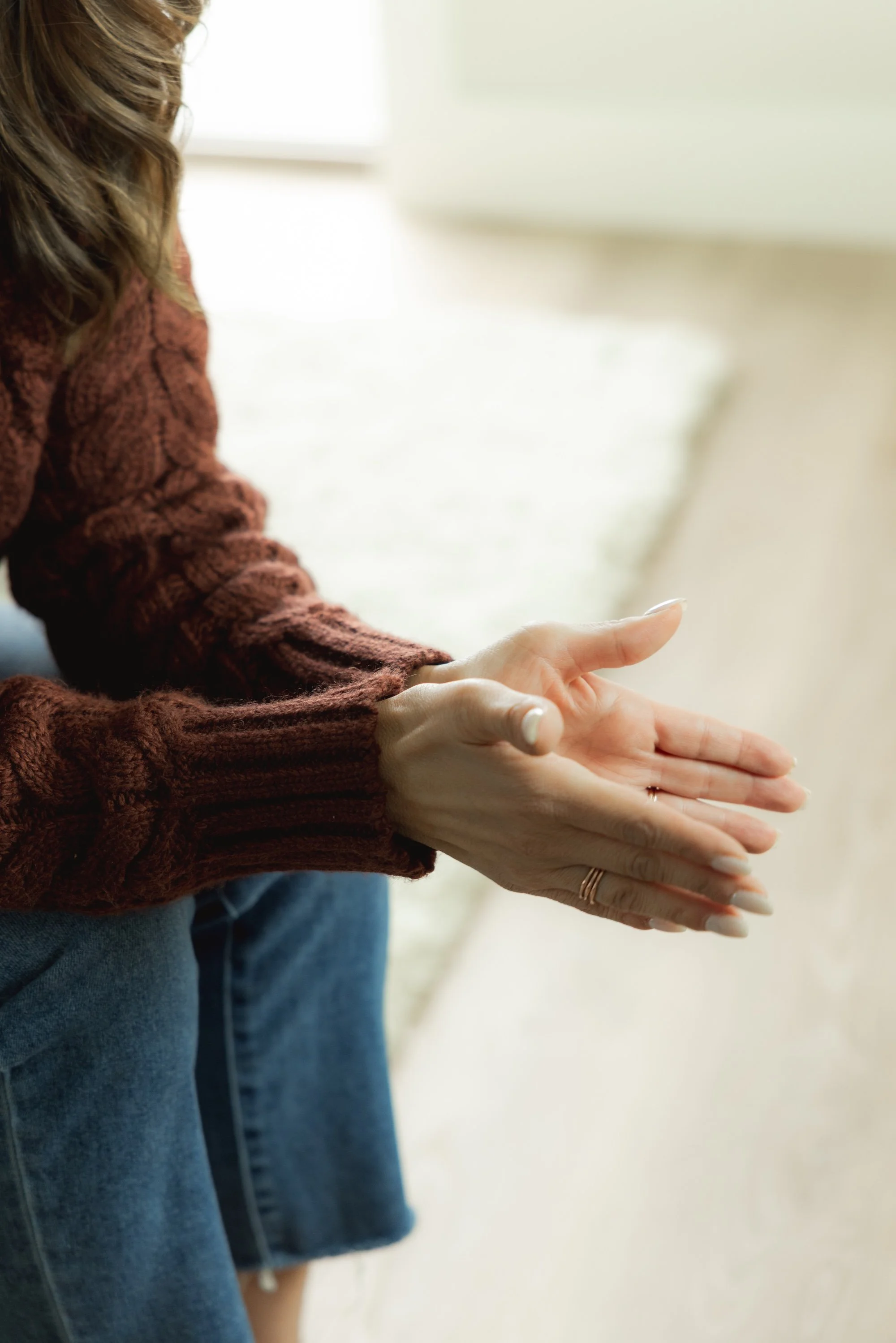 Person wearing a brown sweater sitting with hands clasped together, resting on knees, near a window.