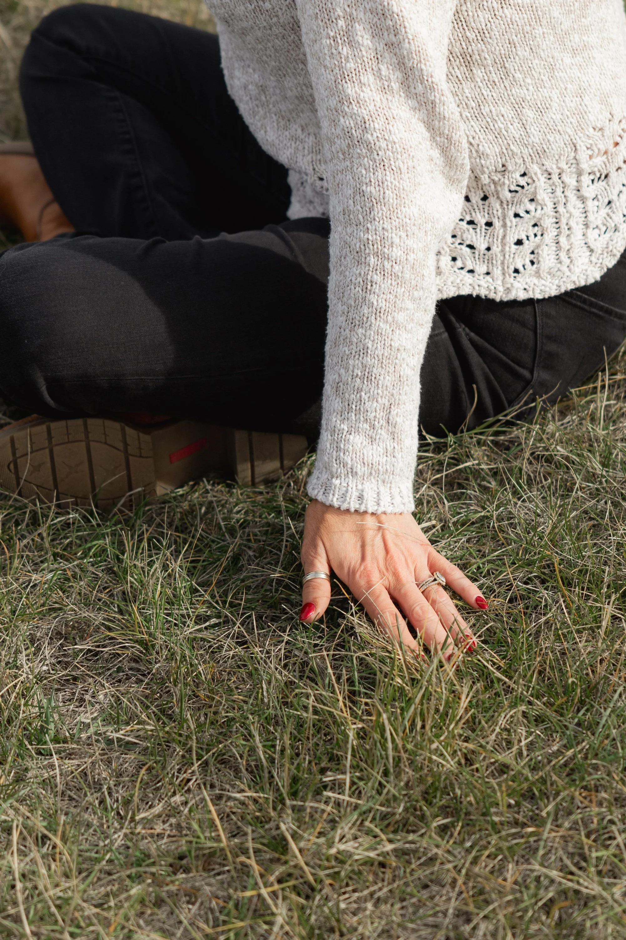 A person sitting on grass outdoors, wearing a cream sweater, black pants, and brown boots, with one hand on the ground and nails painted red.