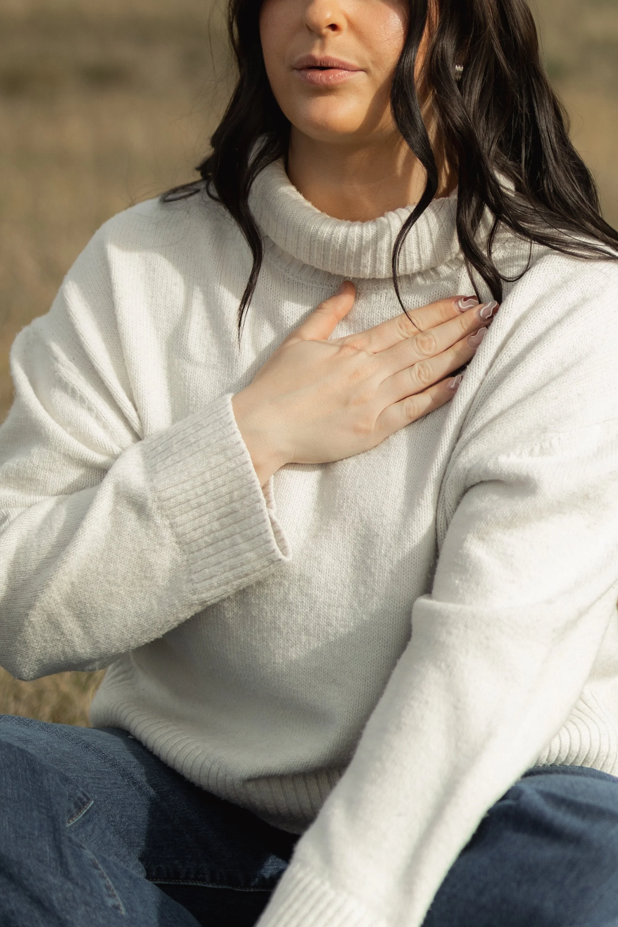 A woman with dark wavy hair wearing a white turtleneck sweater sitting outdoors with her hand over her chest.
