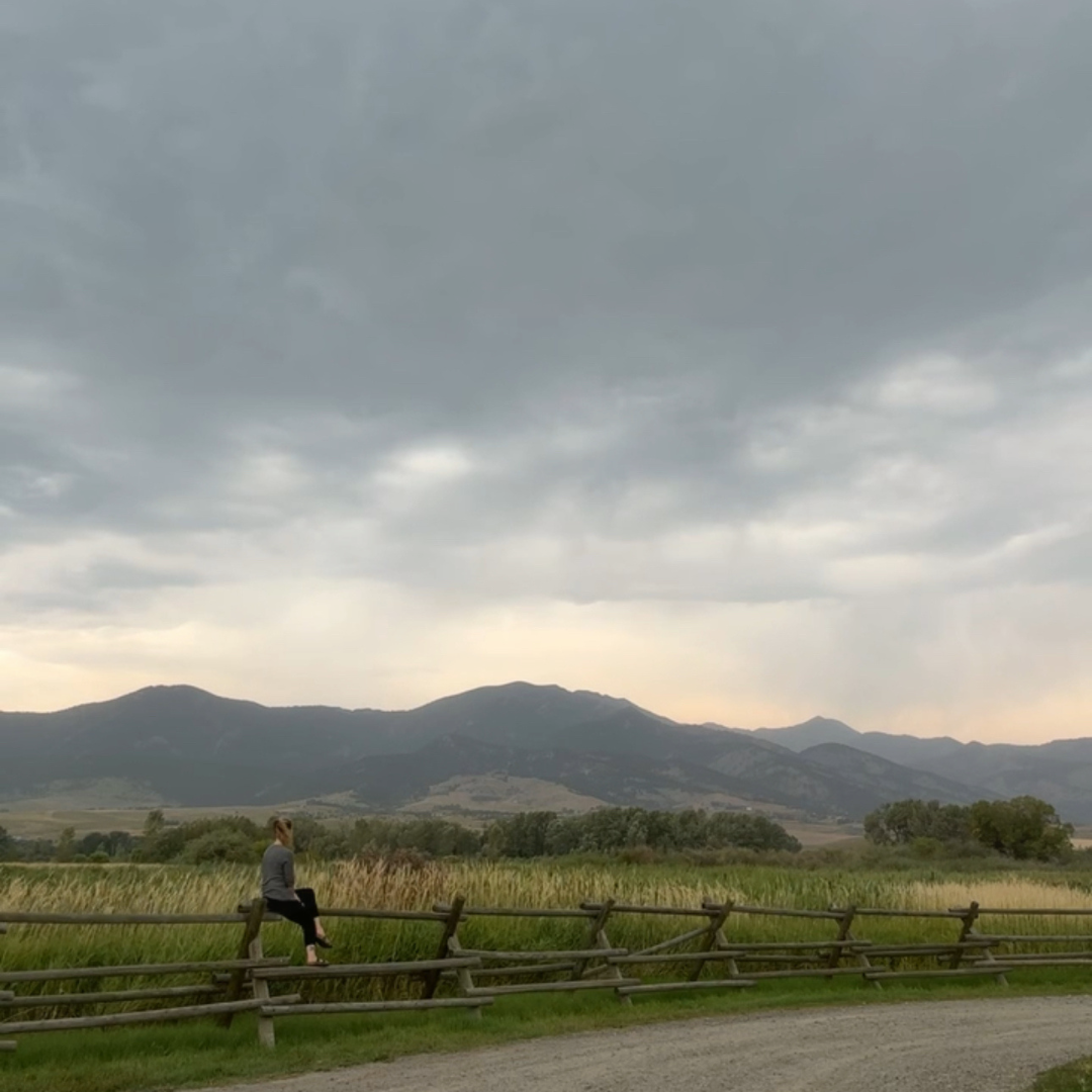 A woman sitting alone on a fence overlooking a vast Montana landscape — a moment of stillness and reflection during burnout recovery