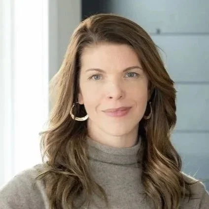 A woman with shoulder-length brown hair, wearing hoop earrings and a light gray turtleneck sweater, standing indoors near a window.