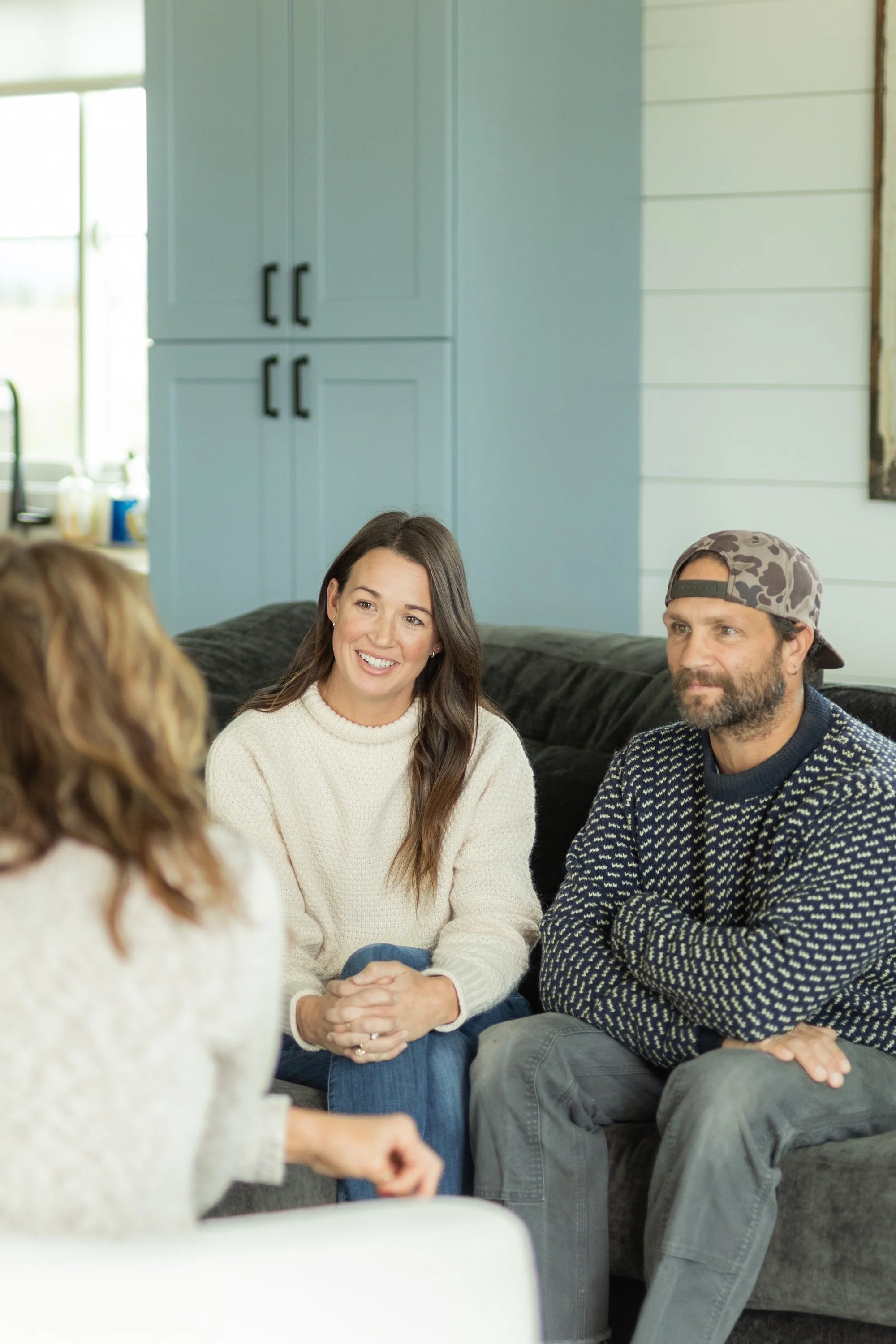Three people sitting on a dark gray couch and talking in a cozy living room with blue walls and a large window in the background.