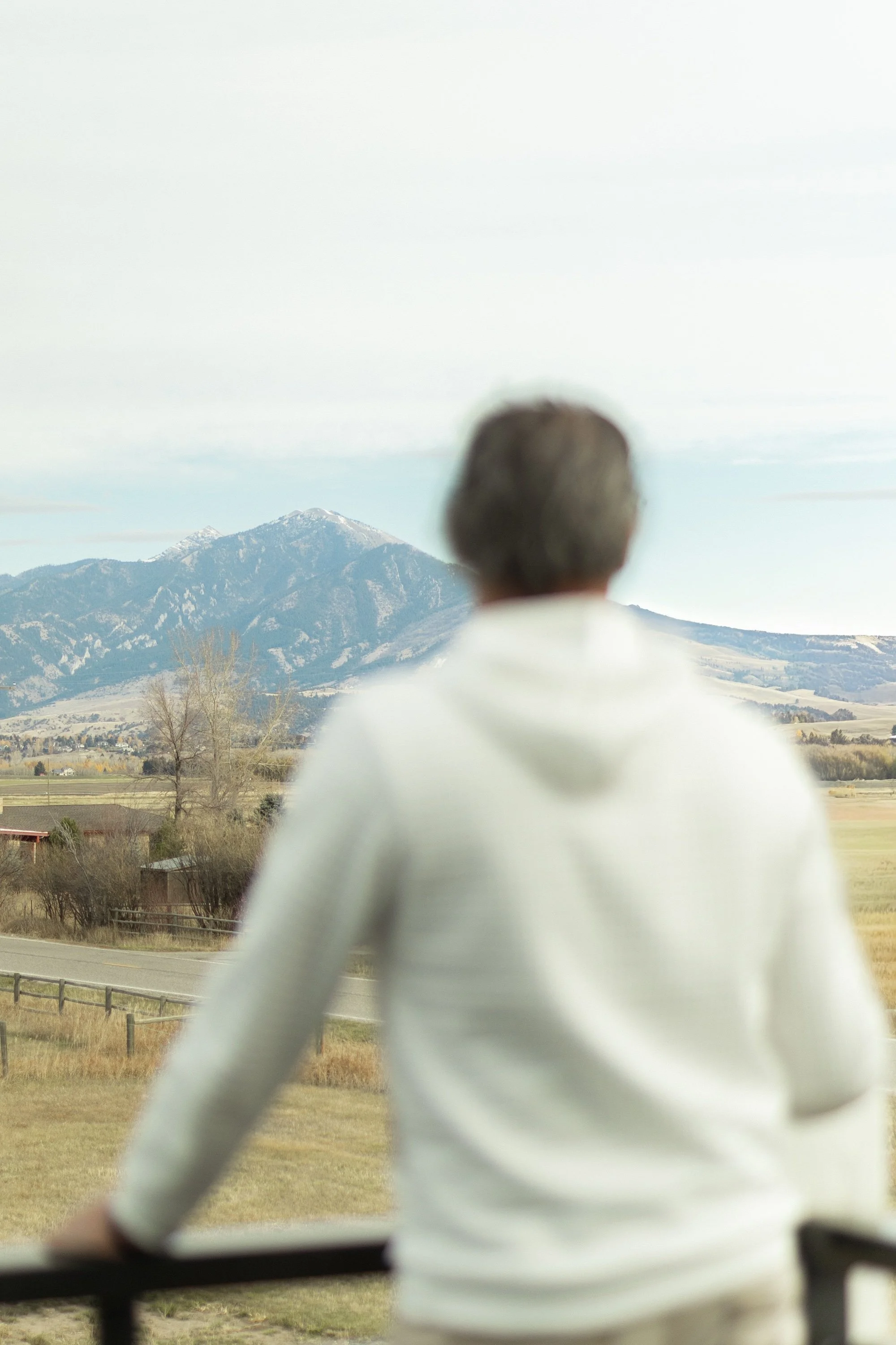 A person in a white hoodie stands outdoors, looking at a mountain range with snow-capped peaks in the distance.