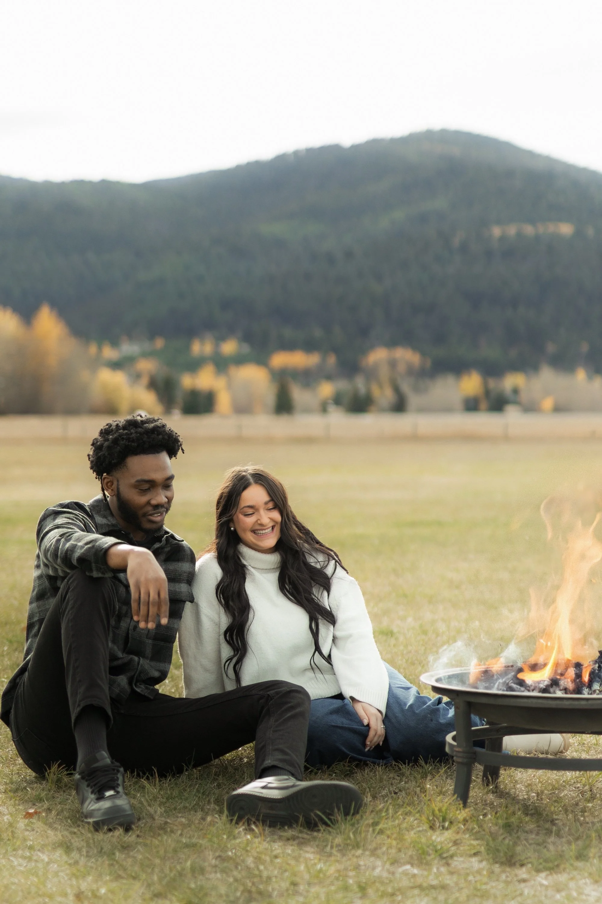 A man and woman sitting on the grass by a campfire outdoors, with mountains in the background.