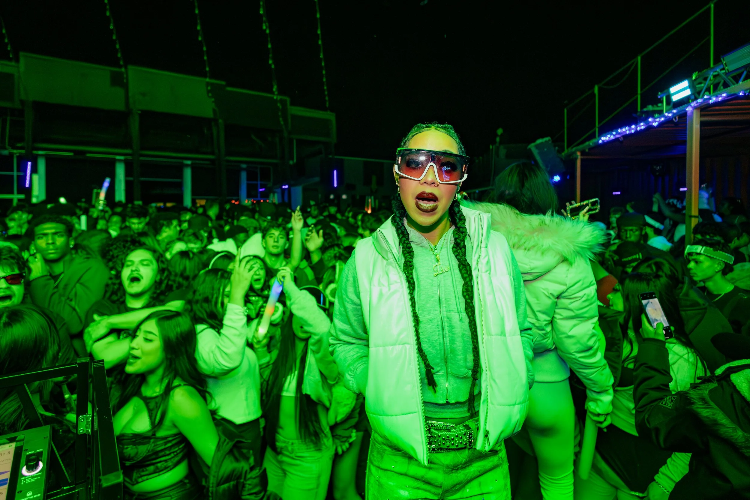 A woman wearing large sunglasses and a puffy jacket stands in a crowded nightclub with green lighting.